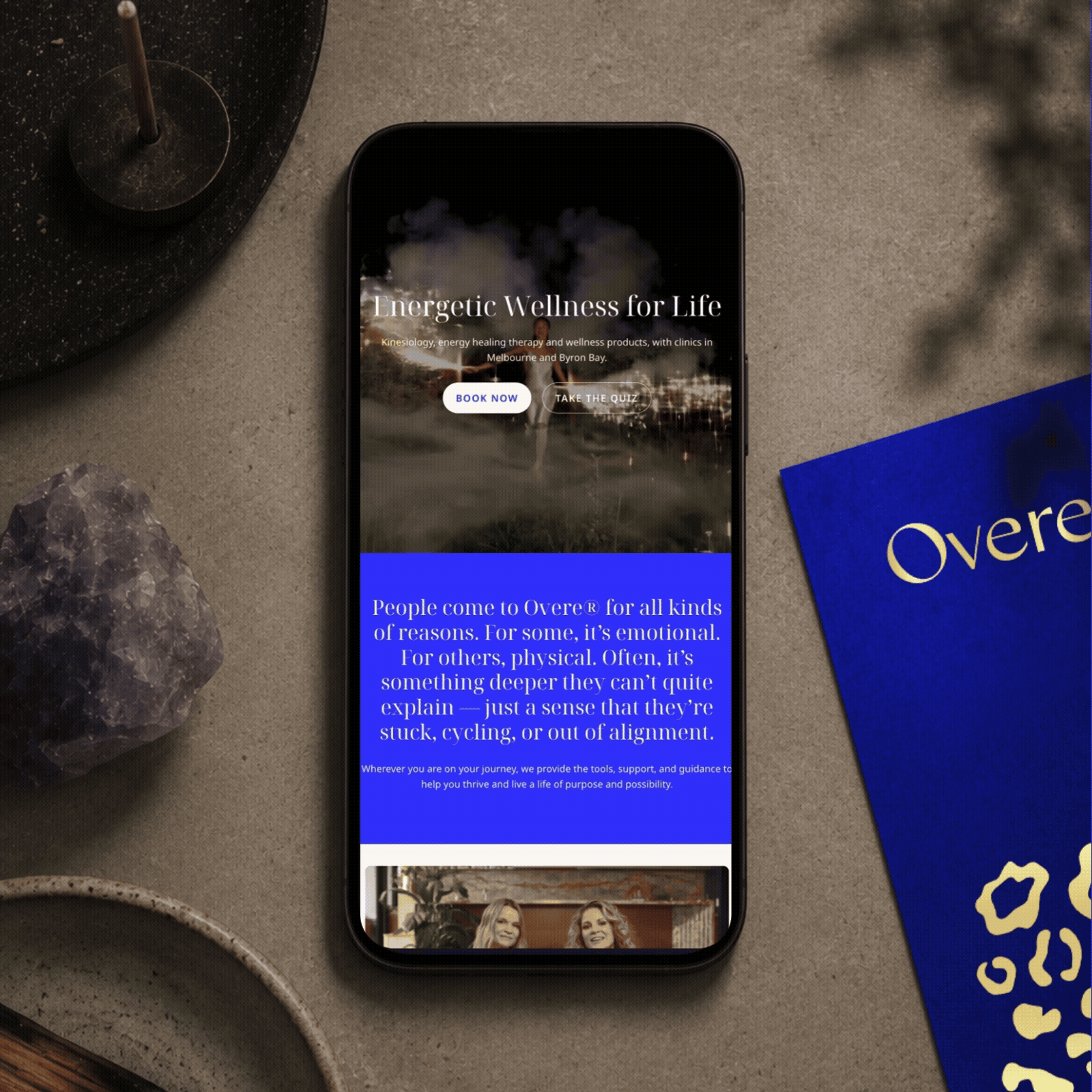 A smartphone displaying a wellness website with the headline 'Energetic Wellness for Life' and two buttons labeled 'Book Now' and 'Take the Quiz'. The phone is on a textured gray surface, surrounded by a speckled black plate with a tall glass, a purple crystal, and a blue booklet.