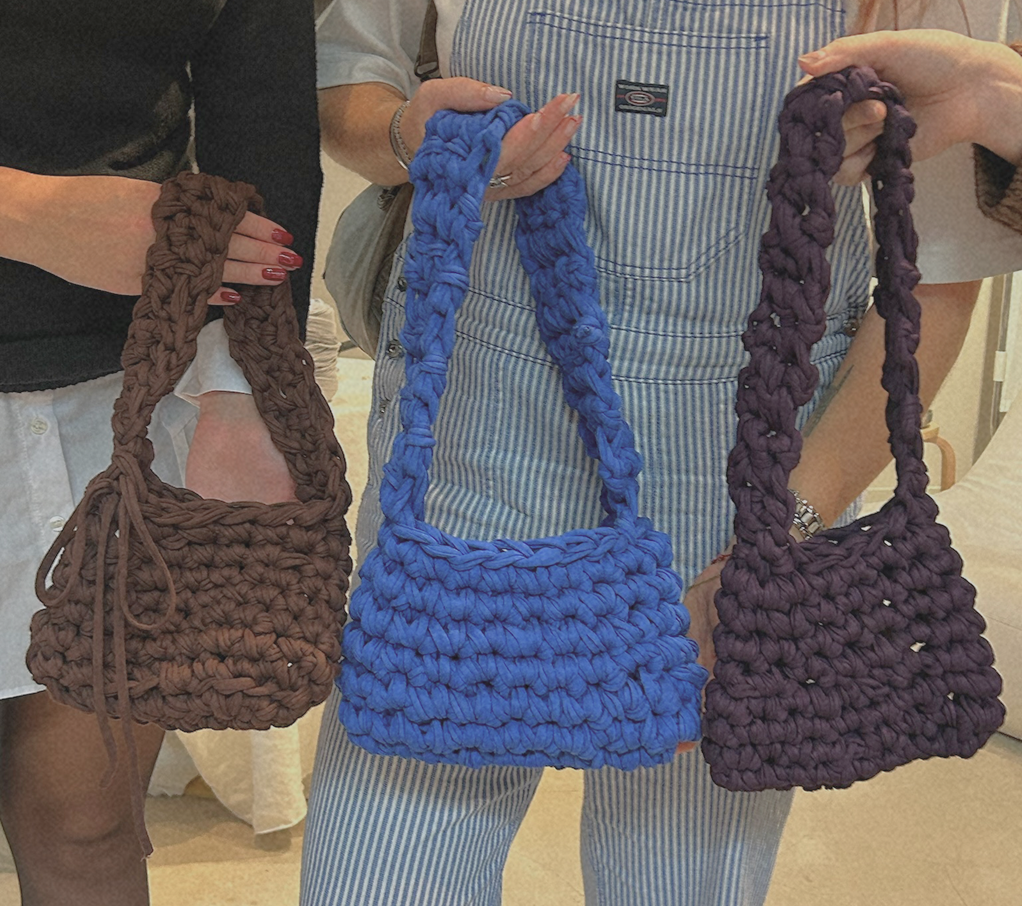 Three women displaying handmade chunky knit bags in brown, blue, and purple in an indoor setting.