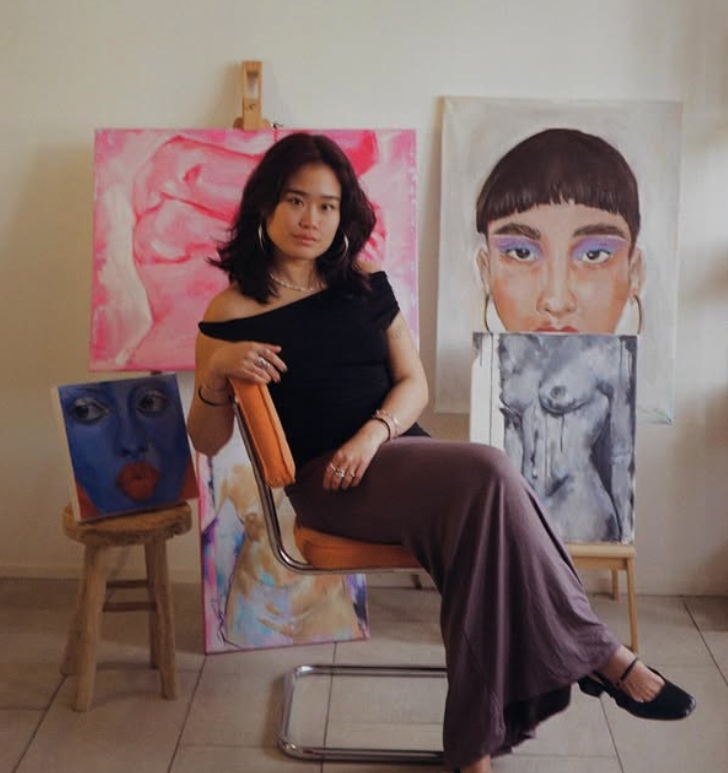 Woman sitting on a chair in front of four colorful paintings, including portraits and abstract art.