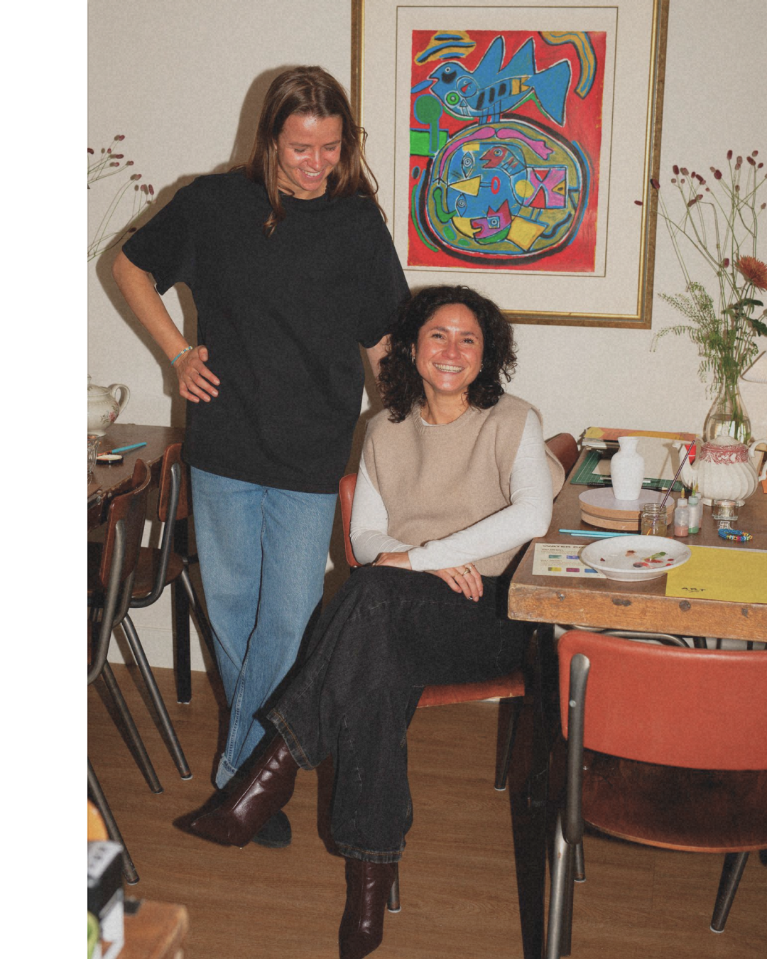 Two women smiling indoors; one stands, the other sits at a table with art supplies, a plate, and flowers; colorful abstract art hangs on the wall behind them.