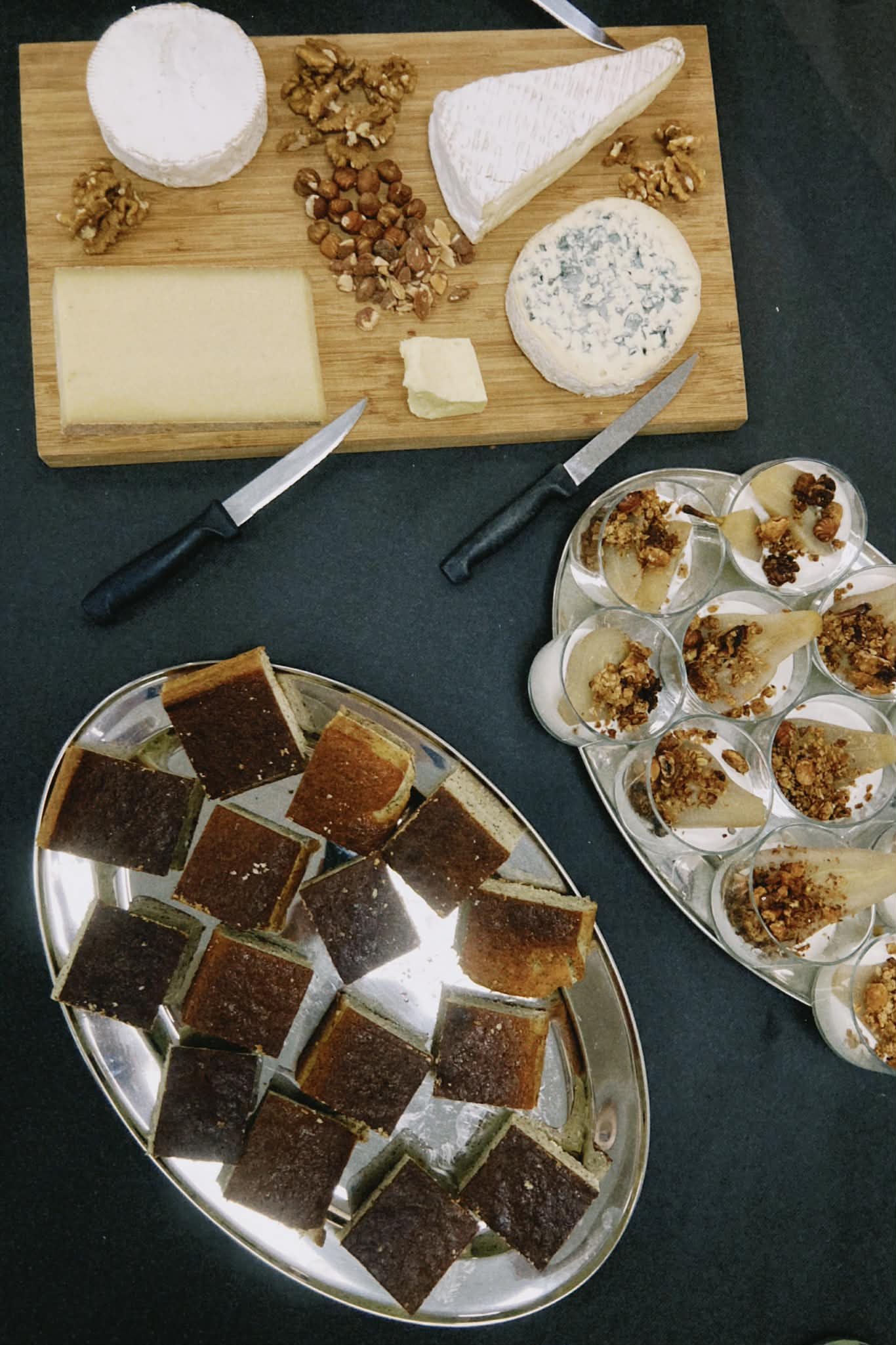 Plateau avec des petits gâteaux carrés, plateau avec des tranches de tarte, plateau avec des verrines de fruits et granola, plateau en bois avec différents types de fromages, noix, noisettes, et une tranche de fromage.