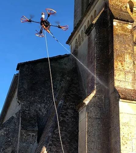 Un drone équipé d'une pompe pulvérise de l'eau sur la façade d'un bâtiment en pierre.