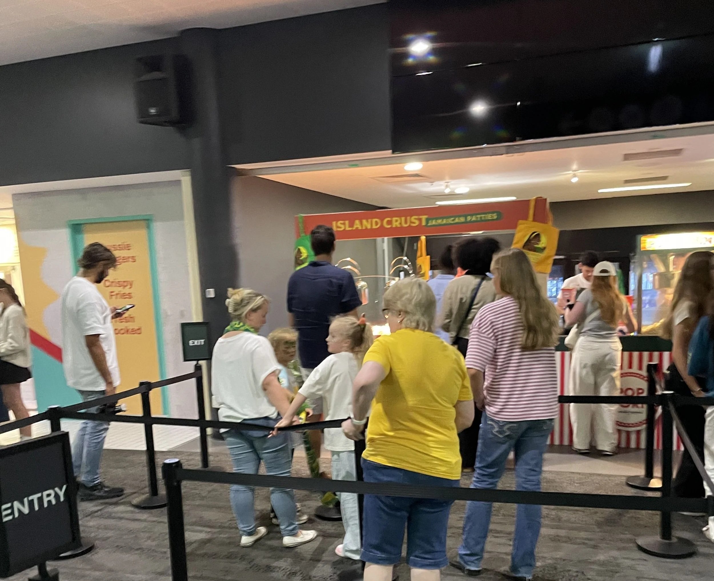People standing in line at a food or snack counter inside a mall or food court, with signs for island crust and crispy fries visible above the counter.