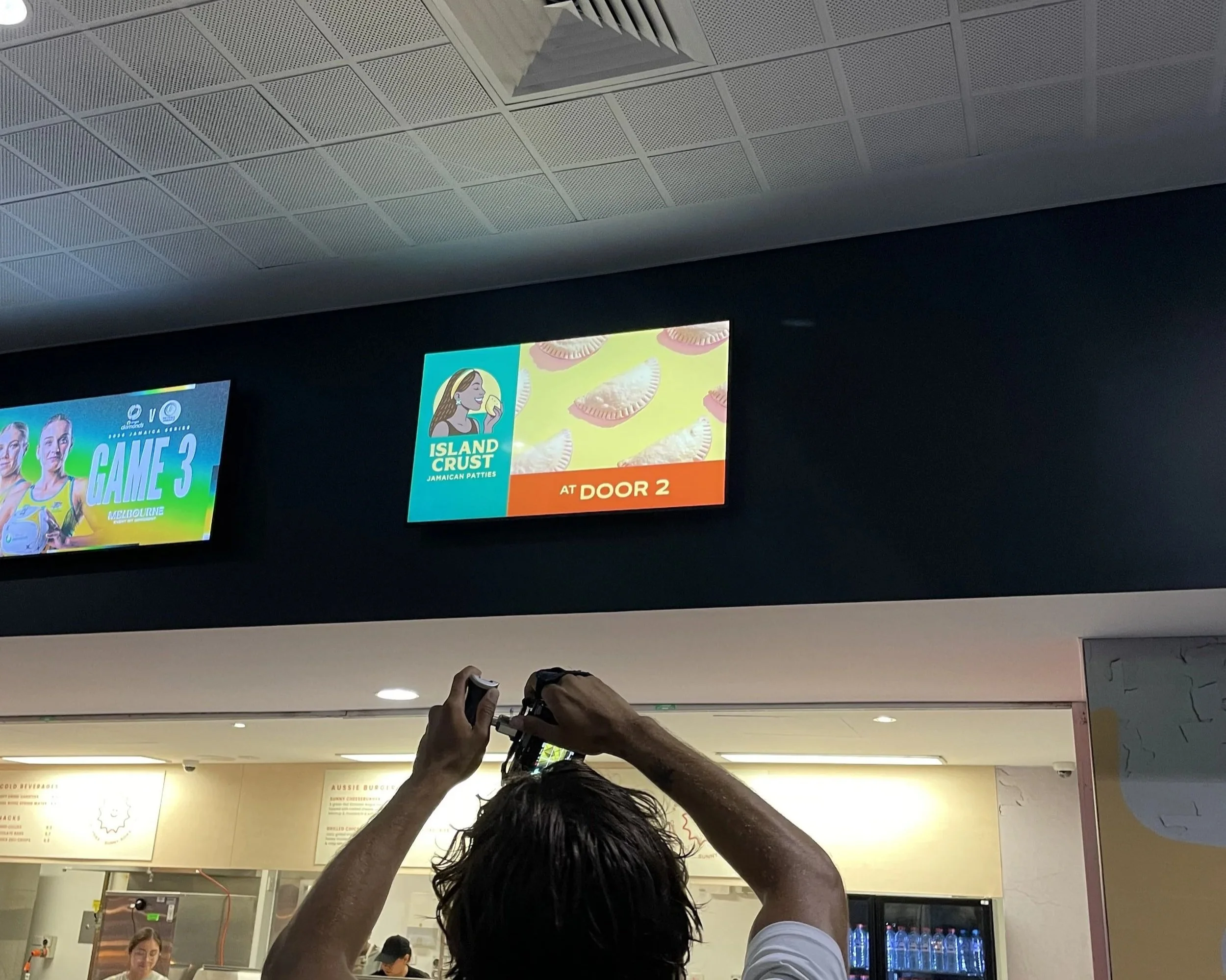 View of a crowded indoor space, likely a restaurant or food court, with two digital signs on the wall. One sign advertises Jamaican patties with an illustration of a woman eating and text, and the other is partially visible. The ceiling has a grid of acoustic panels. Several people are visible, including individuals taking photos with their phones, and a woman in the background appears to be ordering food.