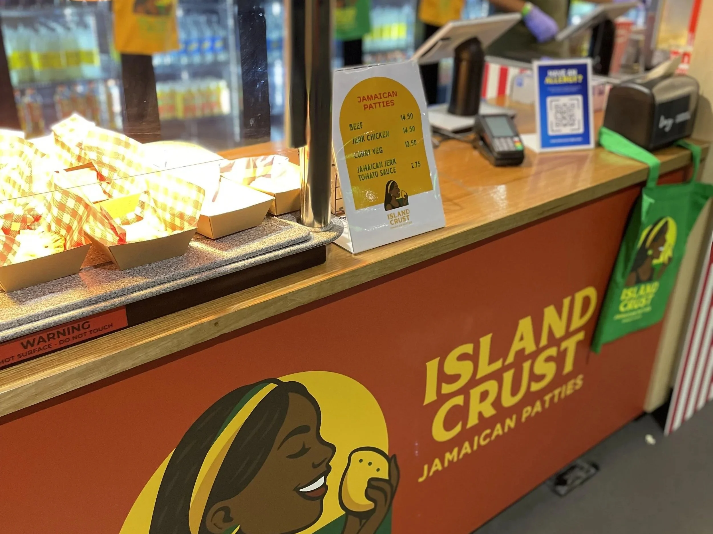 Counter at a store with a sign listing Jamaican patties options including beef, jerk chicken, curry veg, and Jamaican jerk tomato sauce, with the prices. Various products and a point of sale system visible in the background.