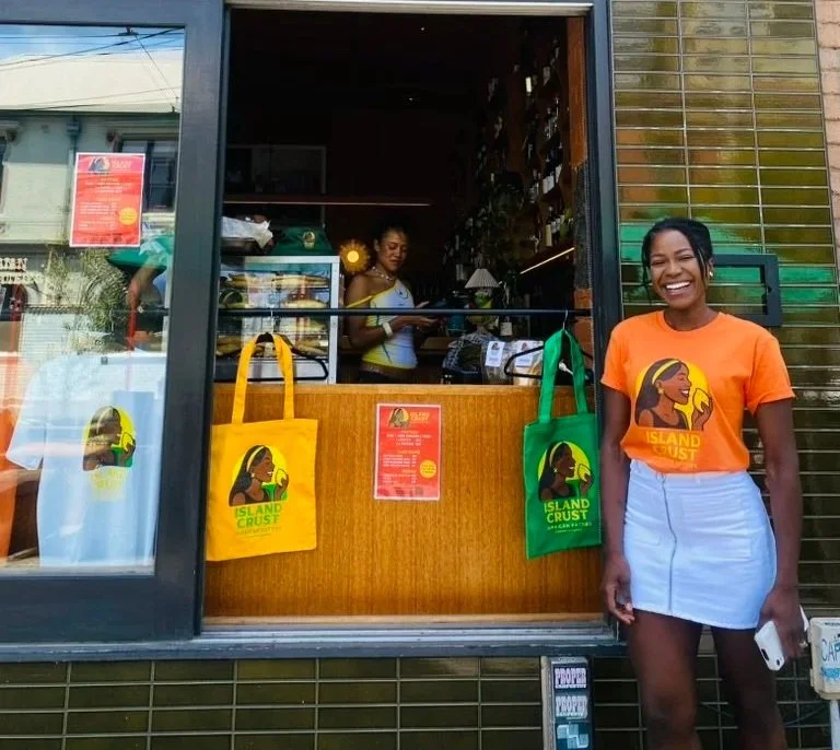 A smiling woman wearing an orange T-shirt and light blue skirt standing outside a small storefront, with two tote bags hanging in the window displaying a logo for Island Crust. Inside, a person is seen behind the counter.