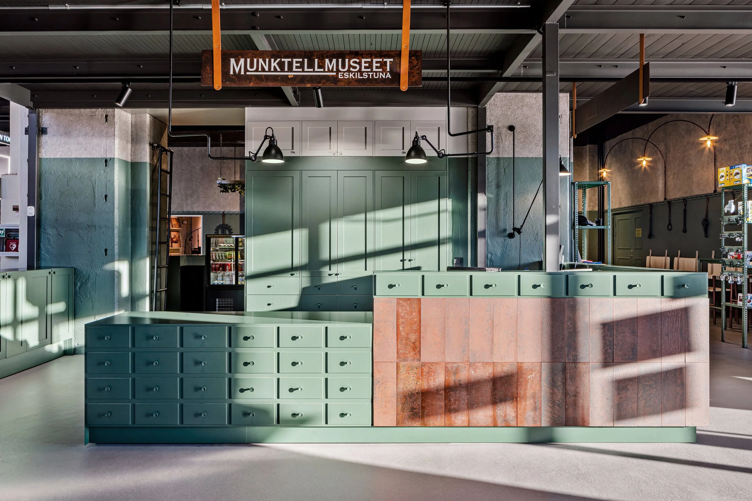 Interior of a store or cafe with a green and brown reception desk, a green cabinet, and a sign that reads 'MUNKTELL MUSSET ESKILSTUNA' hanging from the ceiling.