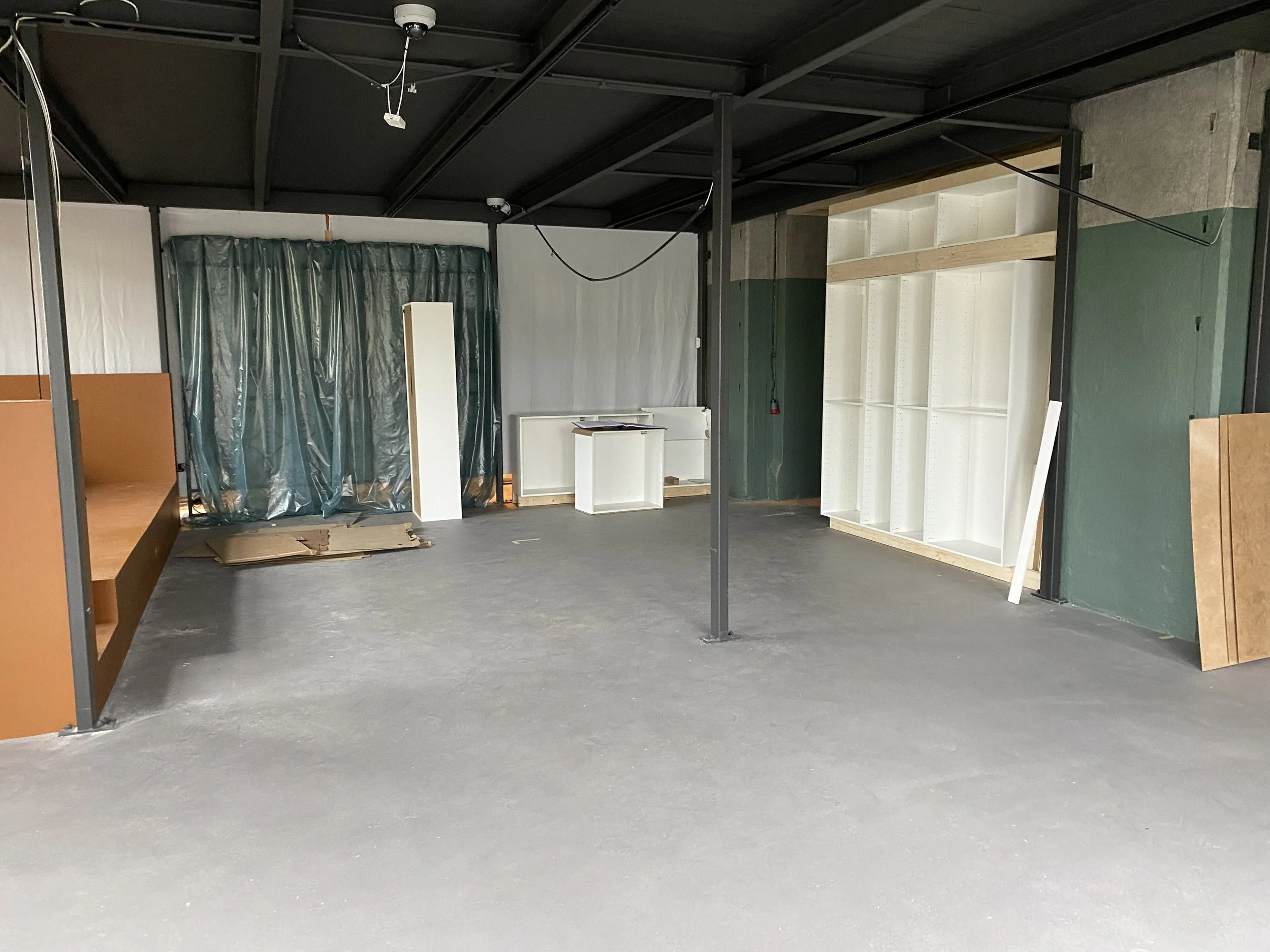 An unfinished retail or storage space with a concrete floor, a black steel ceiling, and partial walls. There is a wooden shelving unit on the right side, a small white desk or counter in the center, and a large white shelving unit on the far right. Green plastic sheeting covers a section at the back, and construction materials and tools are scattered around.