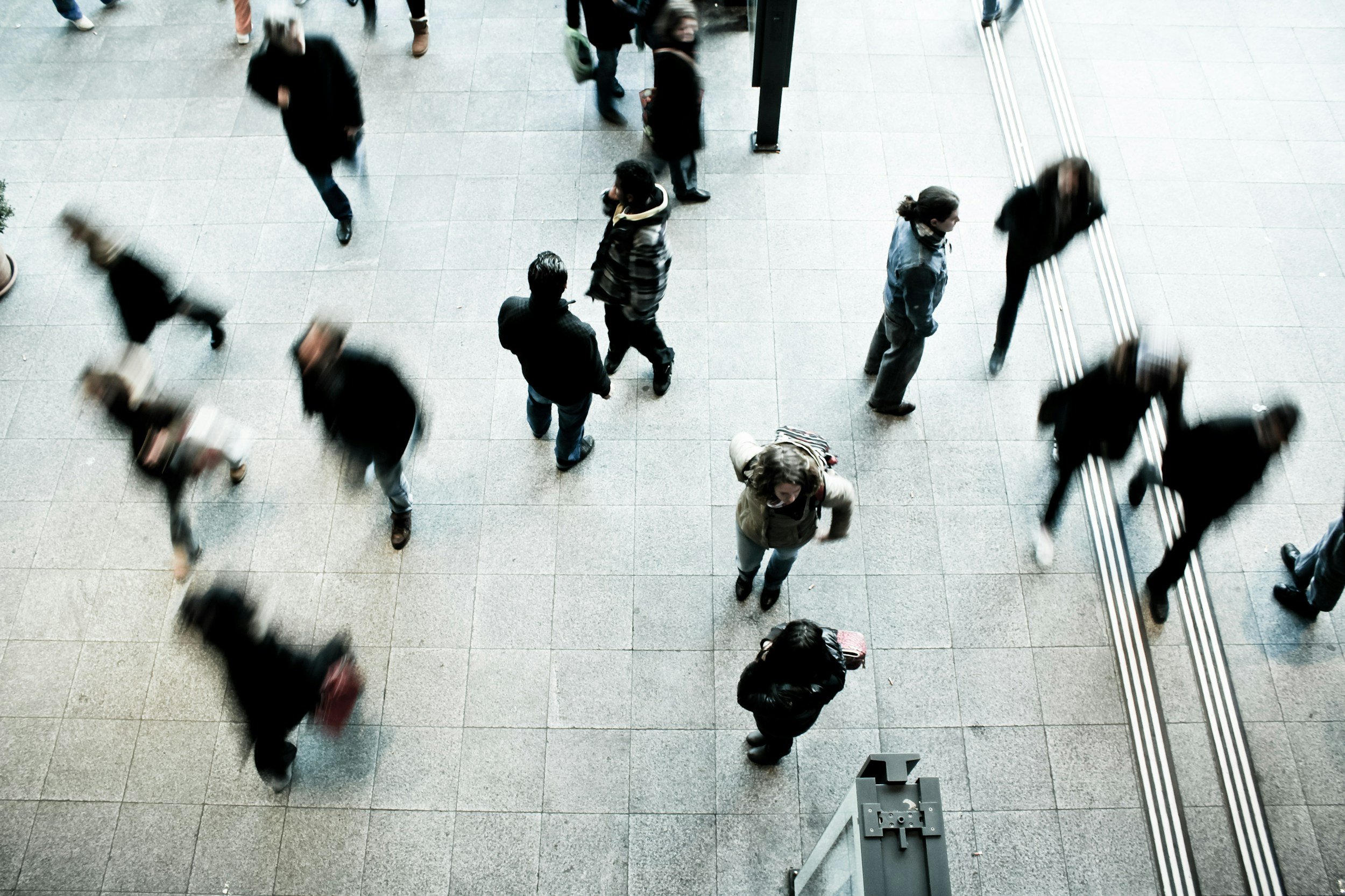People walking on a tiled floor in a public indoor space, some carrying bags and wearing coats.
