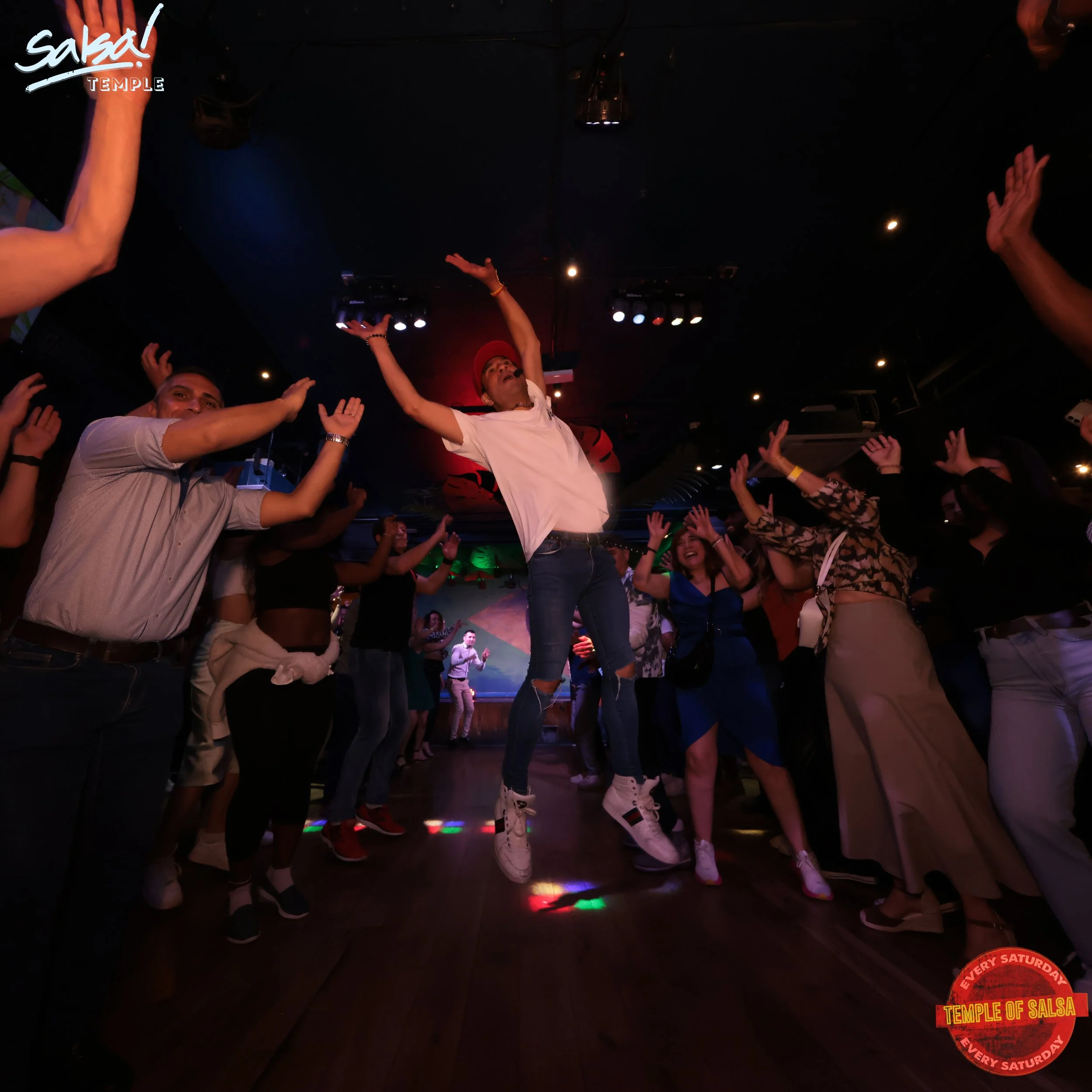 People dancing at a salsa nightclub with colorful lights, with a man jumping in the air in the center, wearing a white t-shirt, ripped jeans, and white sneakers, surrounded by women and men enjoying the music and dance.