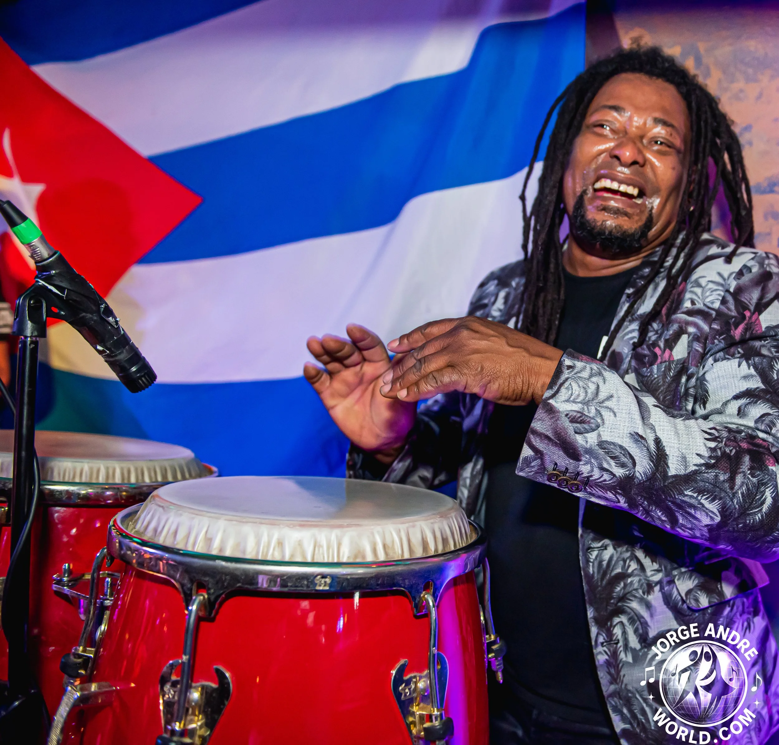 A musician with dreadlocks and a patterned jacket playing congas, with a Cuban flag in the background and a microphone nearby.
