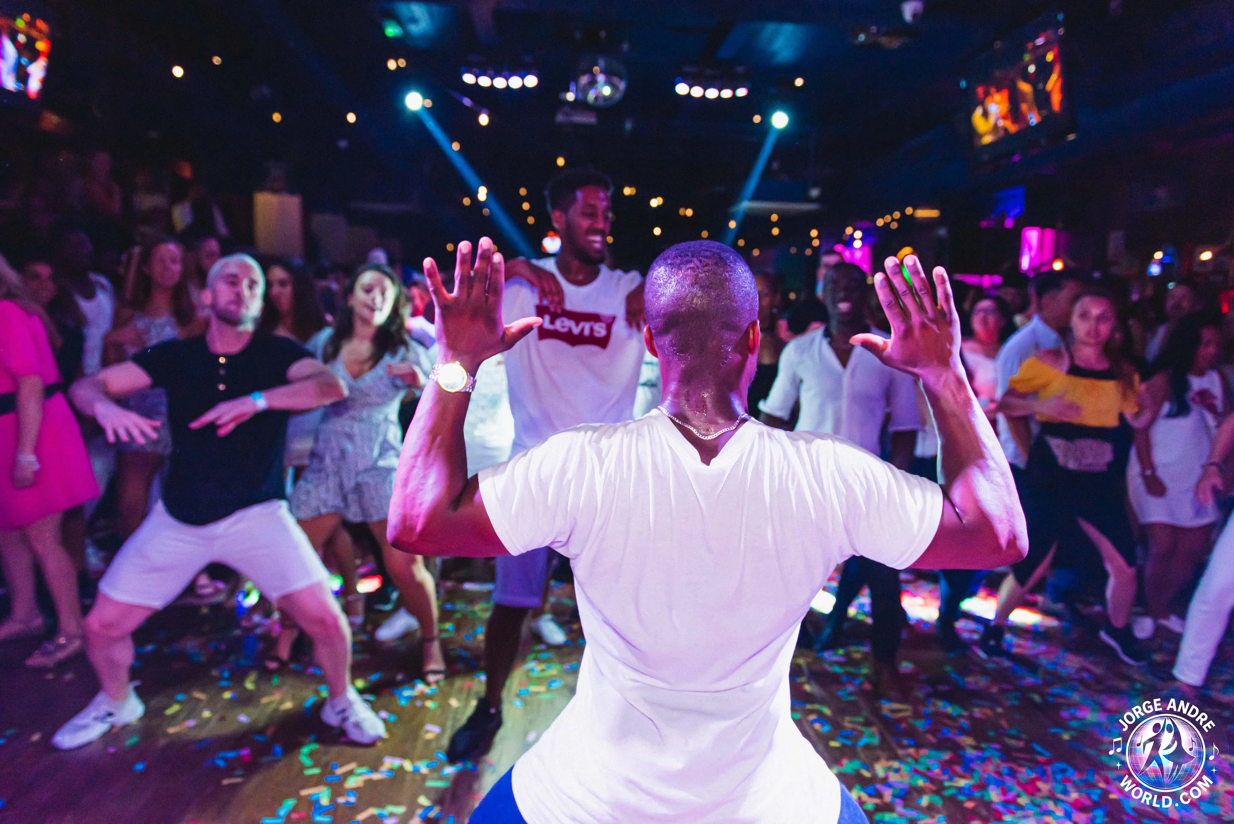 People dancing at a nightclub with colorful lights and confetti on the floor, a man in a white t-shirt with his back facing the camera, and a crowd enjoying the music.