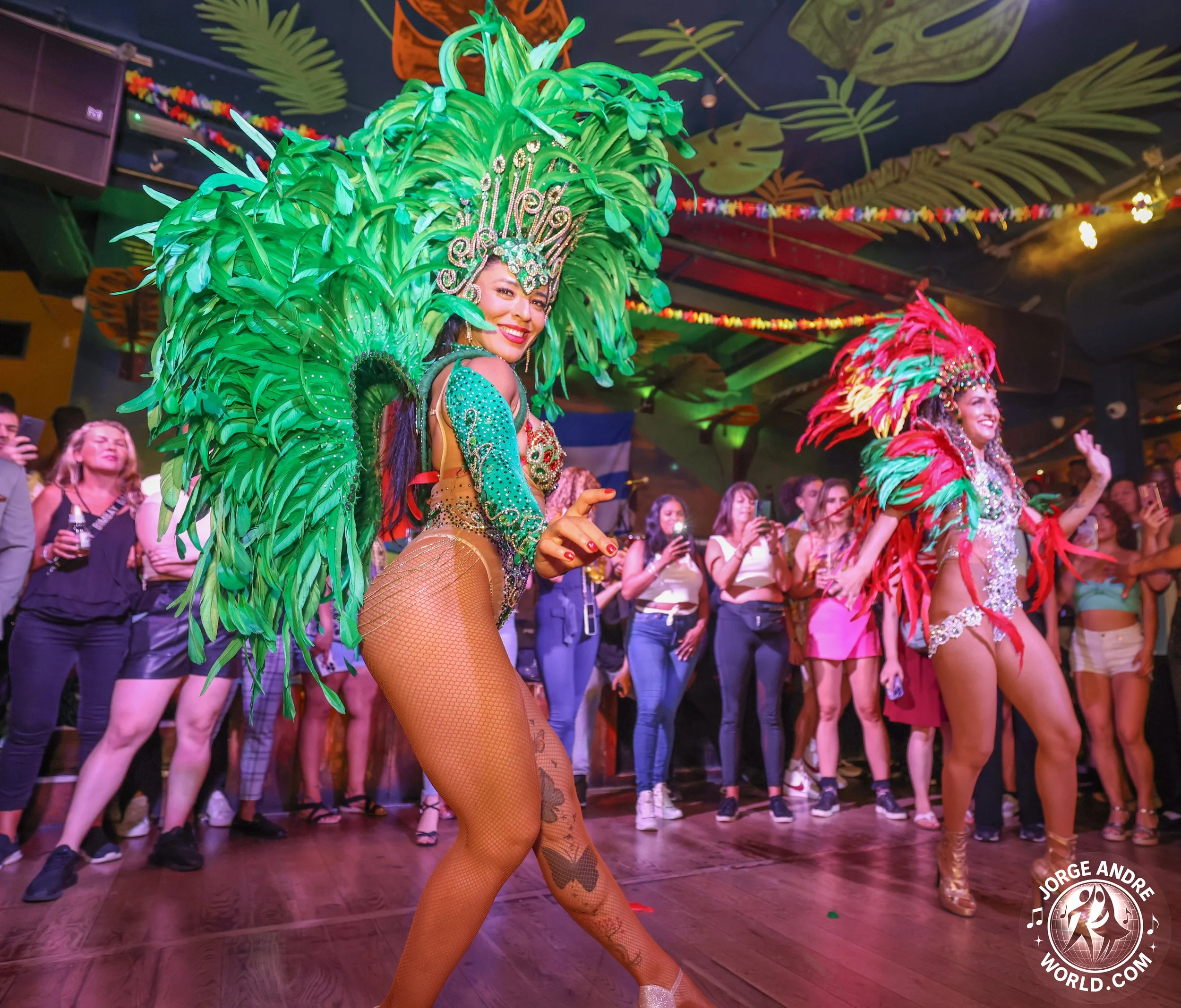 Two women samba dancer dressed in colorful, elaborate samba costumes perform in a lively nightclub with an audience watching and taking photos.