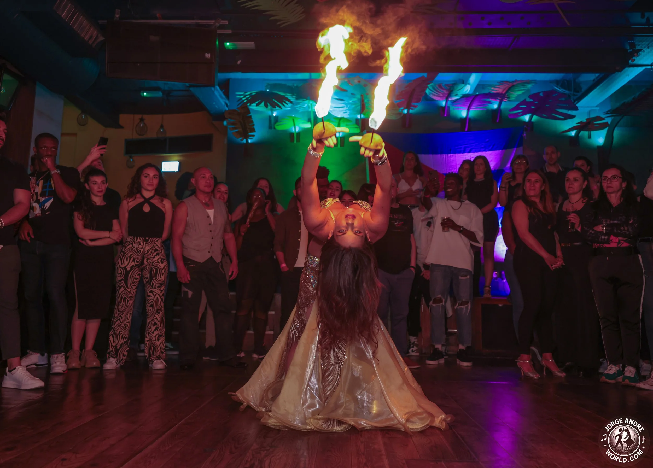 A female performer is upside down on the floor, holding flaming baton wands above her head in a dance routine. She wears a shiny, golden gown, and a crowd of people watches her in a colorful, lively venue with vibrant lighting and tropical decor.