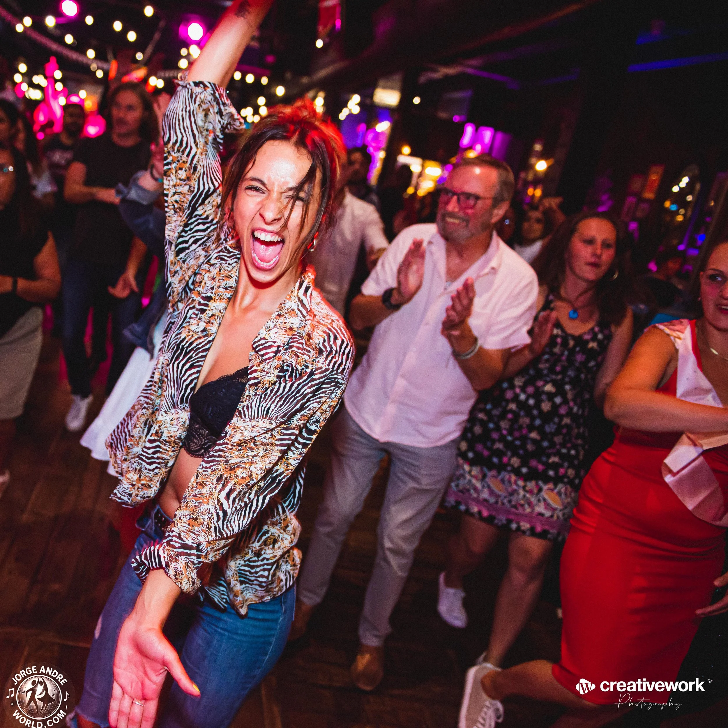 A woman in a zebra-print jacket and jeans dancing and singing with her mouth open, surrounded by people dancing and clapping in a lively party or nightclub setting with colorful lights.