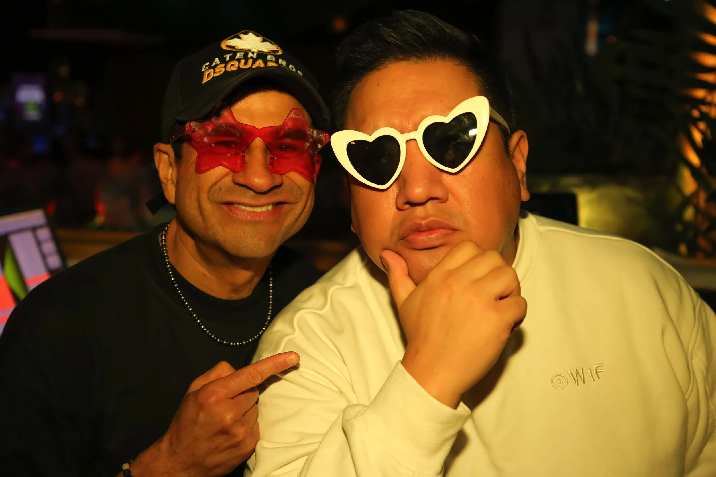 Two men wearing playful sunglasses, one with red star-shaped glasses and a black cap, smiling and pointing at the other, who is wearing white heart-shaped glasses and has a contemplative expression.