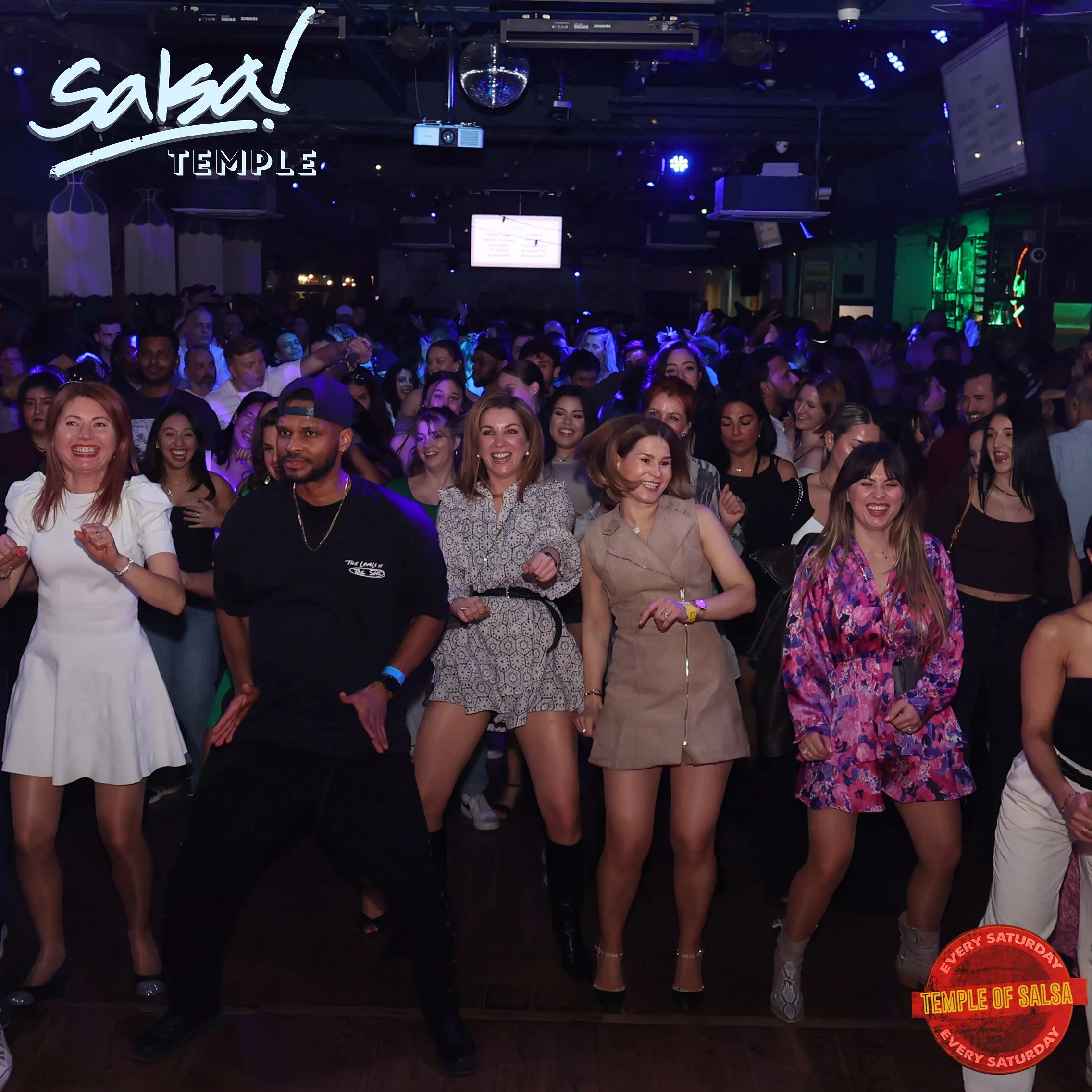 A lively crowd gathered at a salsa dance event called 'Salsa Temple' with some people dancing, smiling, and enjoying the music in a dimly-lit club with colorful lighting and a disco ball overhead.