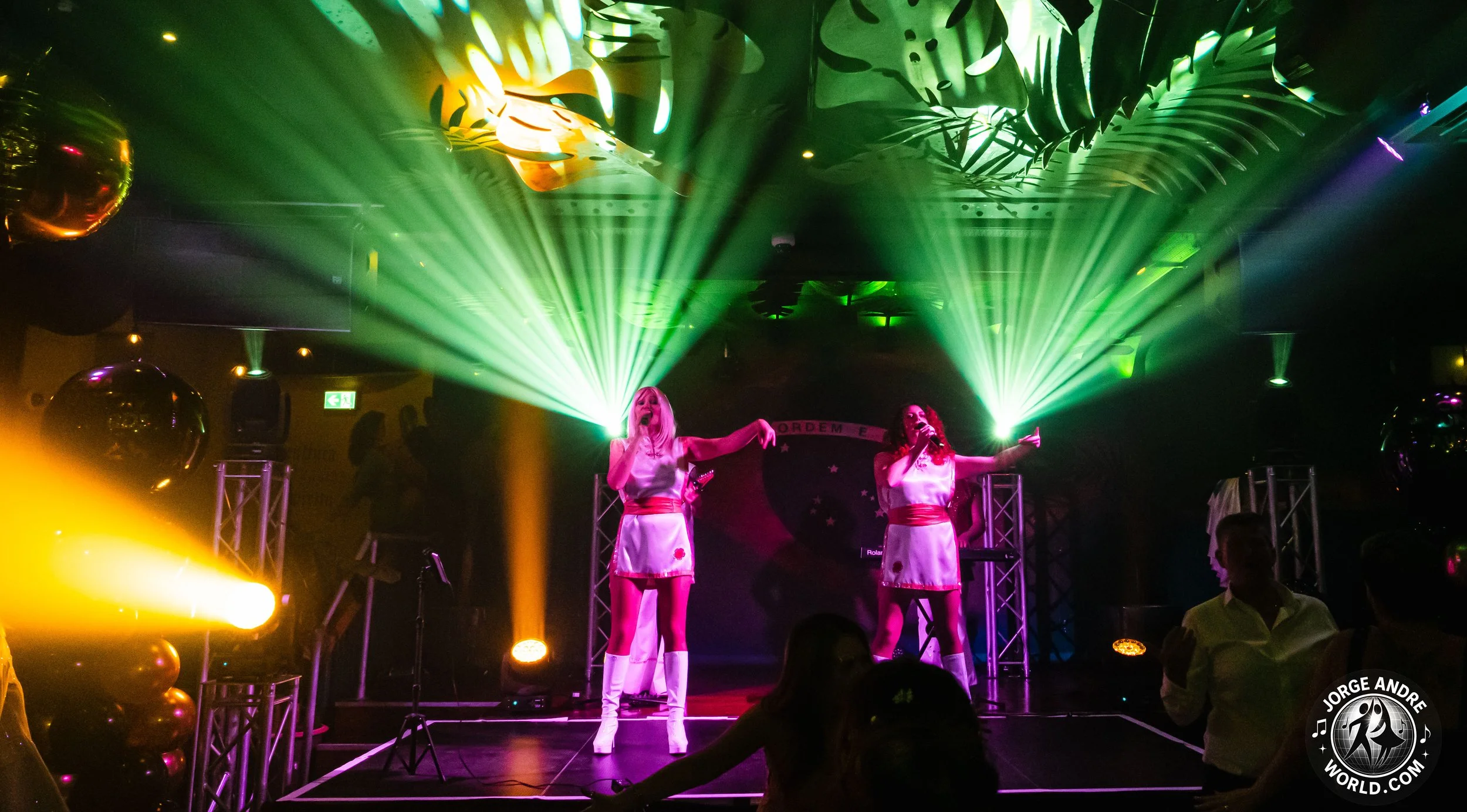 Two female performers on stage singing with a microphone, illuminated by green and yellow stage lights, with an audience in the foreground and decorative balloons and stage equipment around them.
