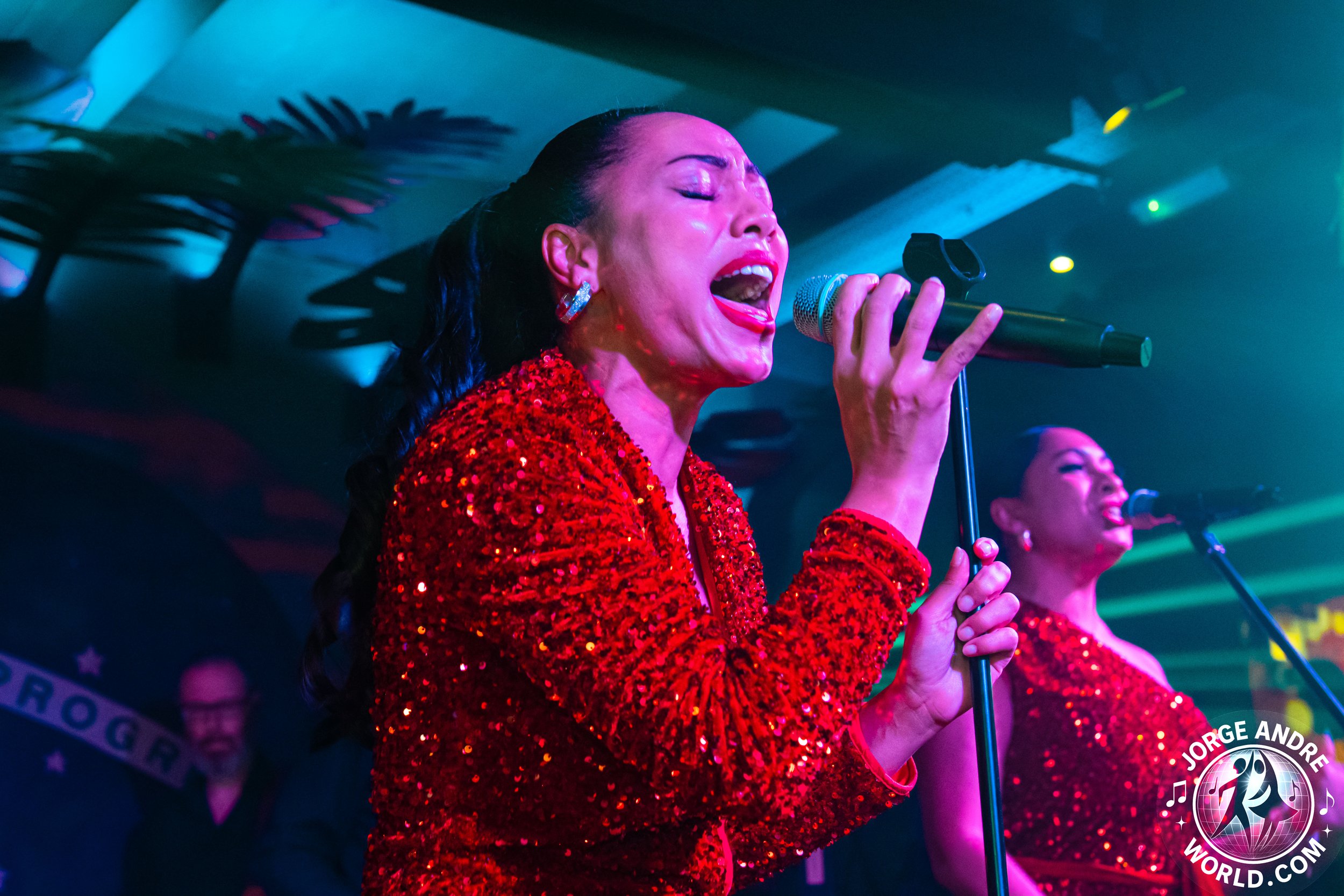 Two women singing passionately into microphones on stage, both wearing shiny red sequined dresses, with a colorful performance lighting and a musician in the background.