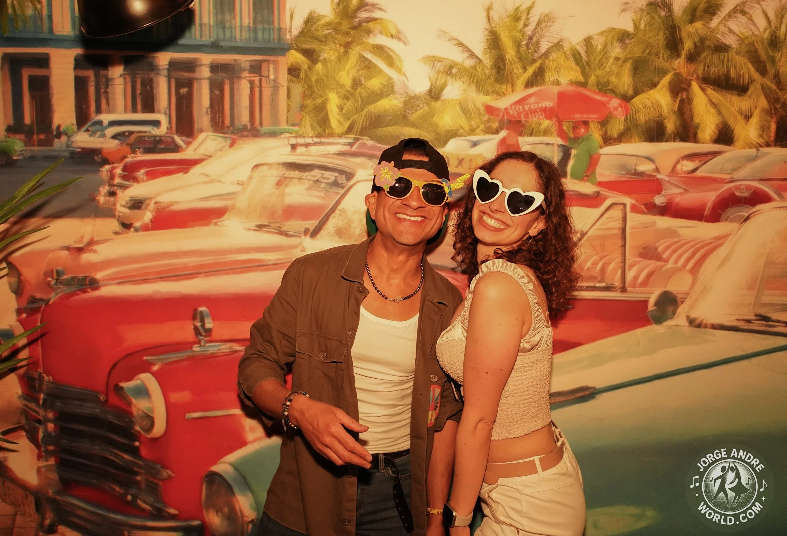 Two people smiling and wearing sunglasses, standing in front of a colorful vintage car backdrop with palm trees and parked cars.