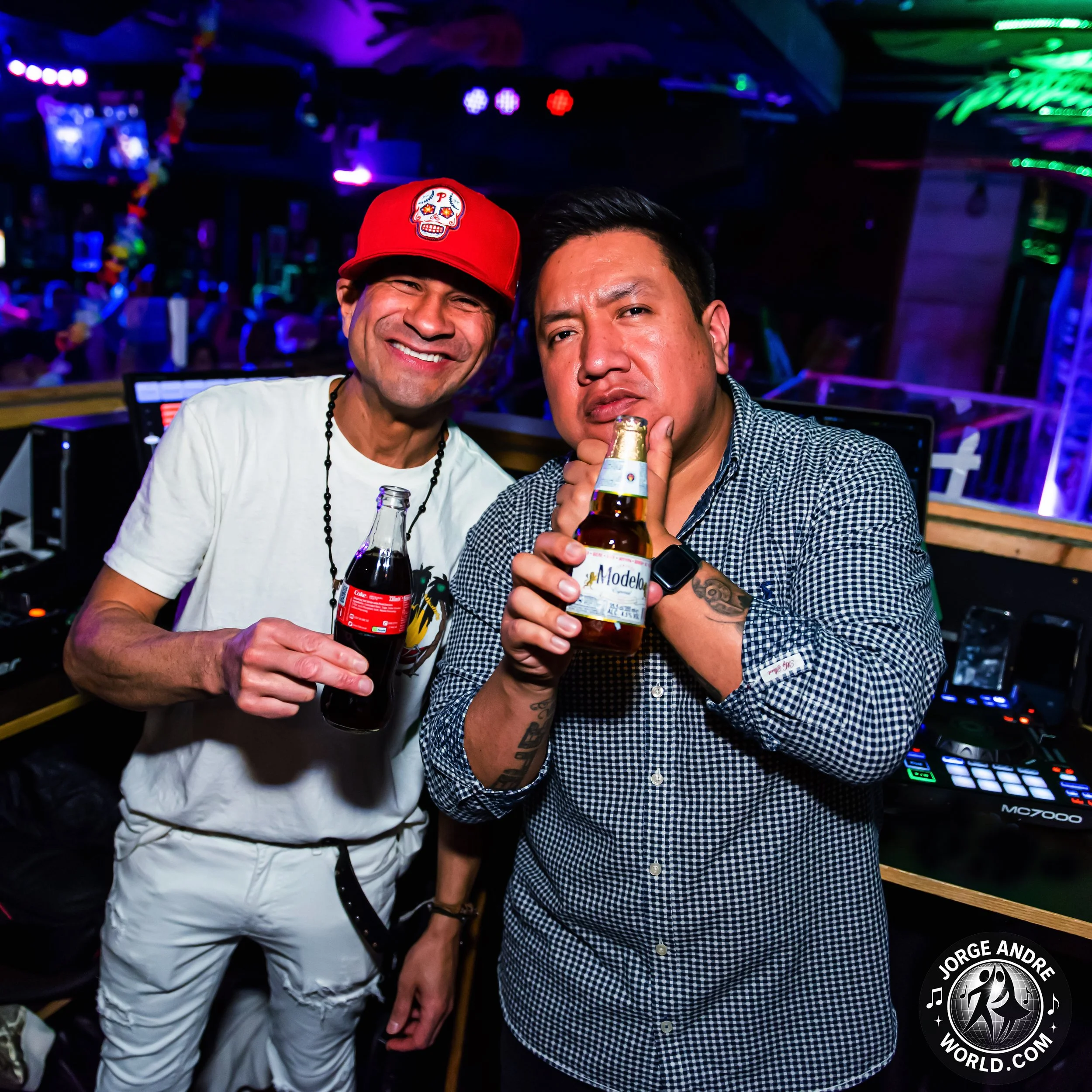 Two men at a nightclub holding drinks, one smiling wearing a red cap with a skull logo and the other with a serious expression in a checkered shirt, with DJ equipment in the background and colorful lighting.