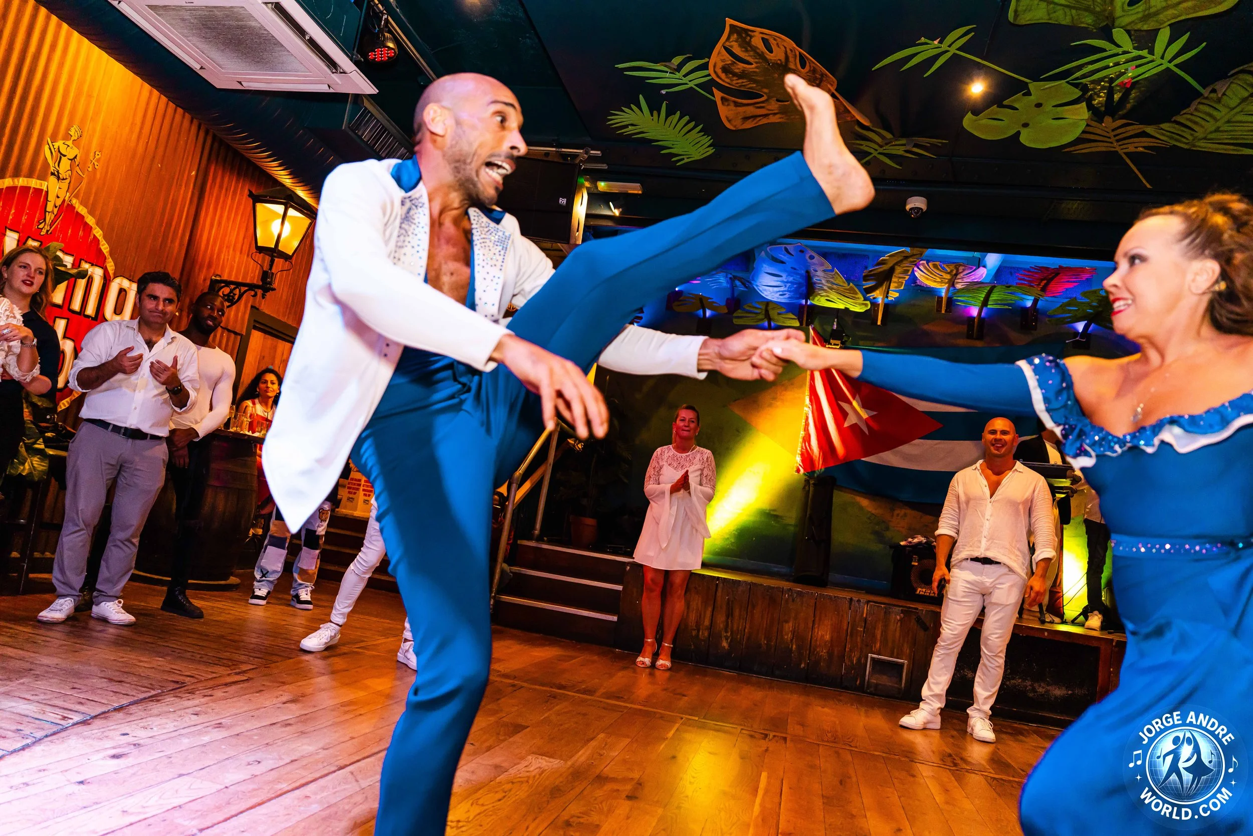 Two dancers in vibrant costumes performing a lively dance in a tropical-themed venue, with onlookers clapping and smiling in the background.