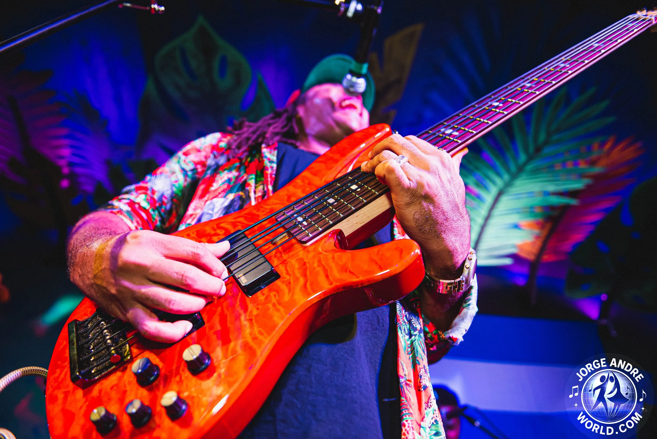 A man playing an orange electric bass guitar on stage with colorful tropical plant decorations in the background.