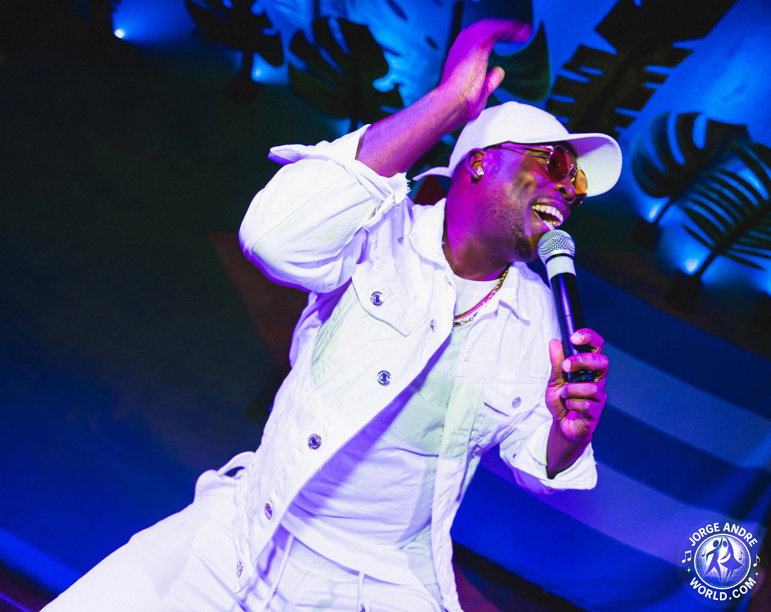 A person wearing white clothing, a white hat, and sunglasses is singing into a microphone on stage. They are smiling and appear to be enjoying the performance, with a blue-lit background.