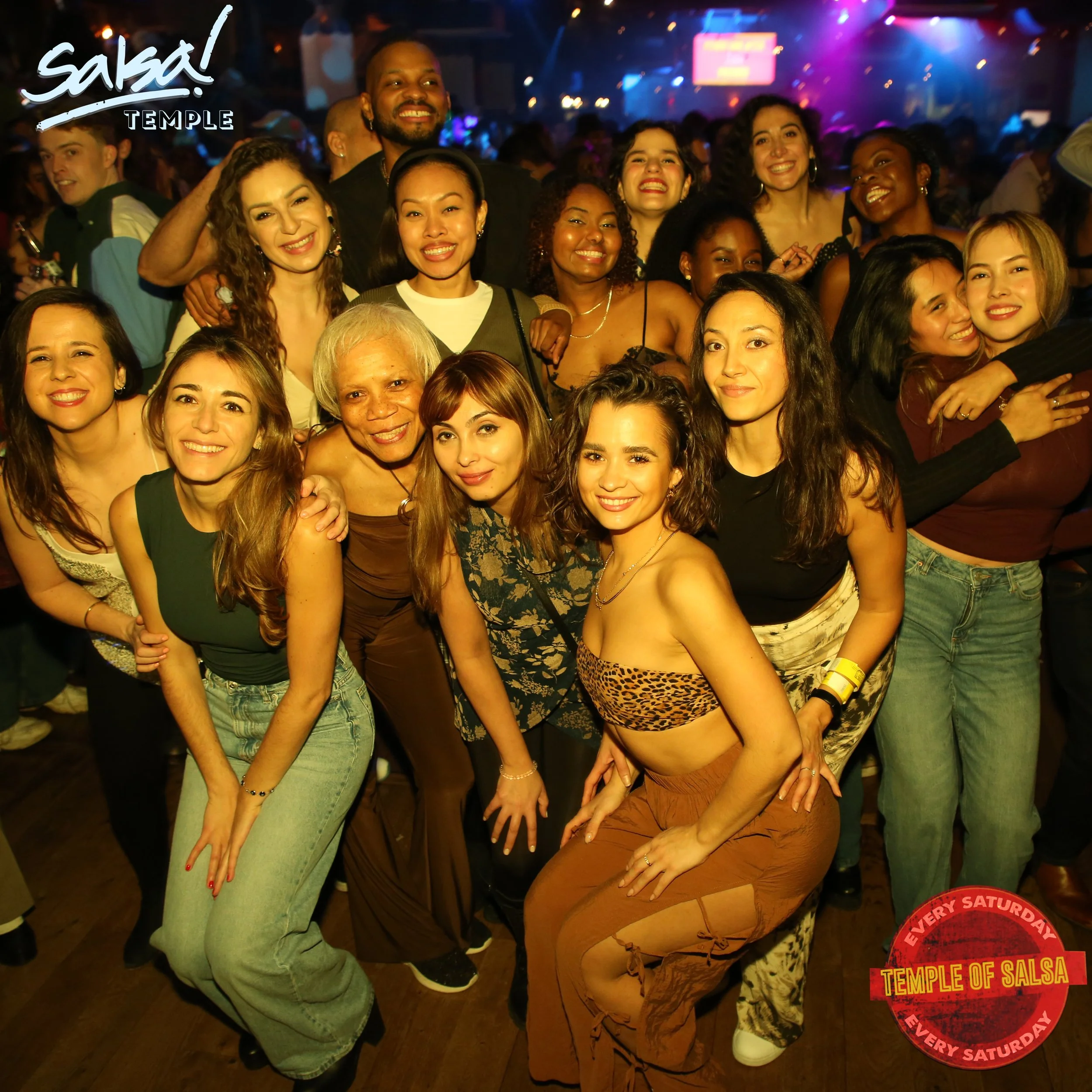 Group of people smiling and posing together at a salsa dance event in a nightclub with colorful lights, with a 'Salsa Temple' logo indicating it occurs every Saturday.