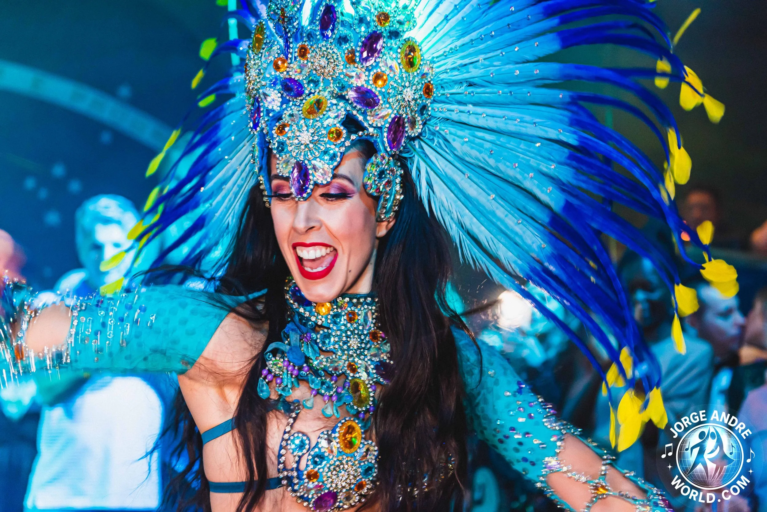 A woman wearing an elaborate blue carnival costume with a large feathered headdress decorated with colorful jewels and sequins, smiling with red lipstick, at a festive event.