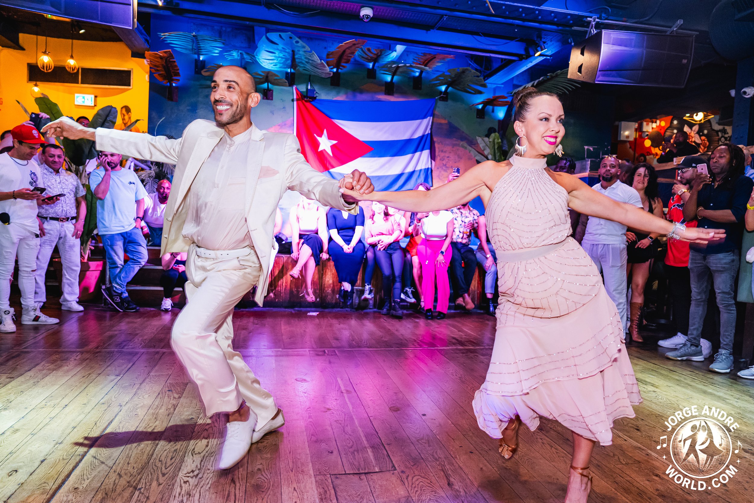 A man and woman dancing together in a lively venue with a crowd watching. The man is dressed in white and the woman in a light pink dress. Cuban flag is visible in the background.