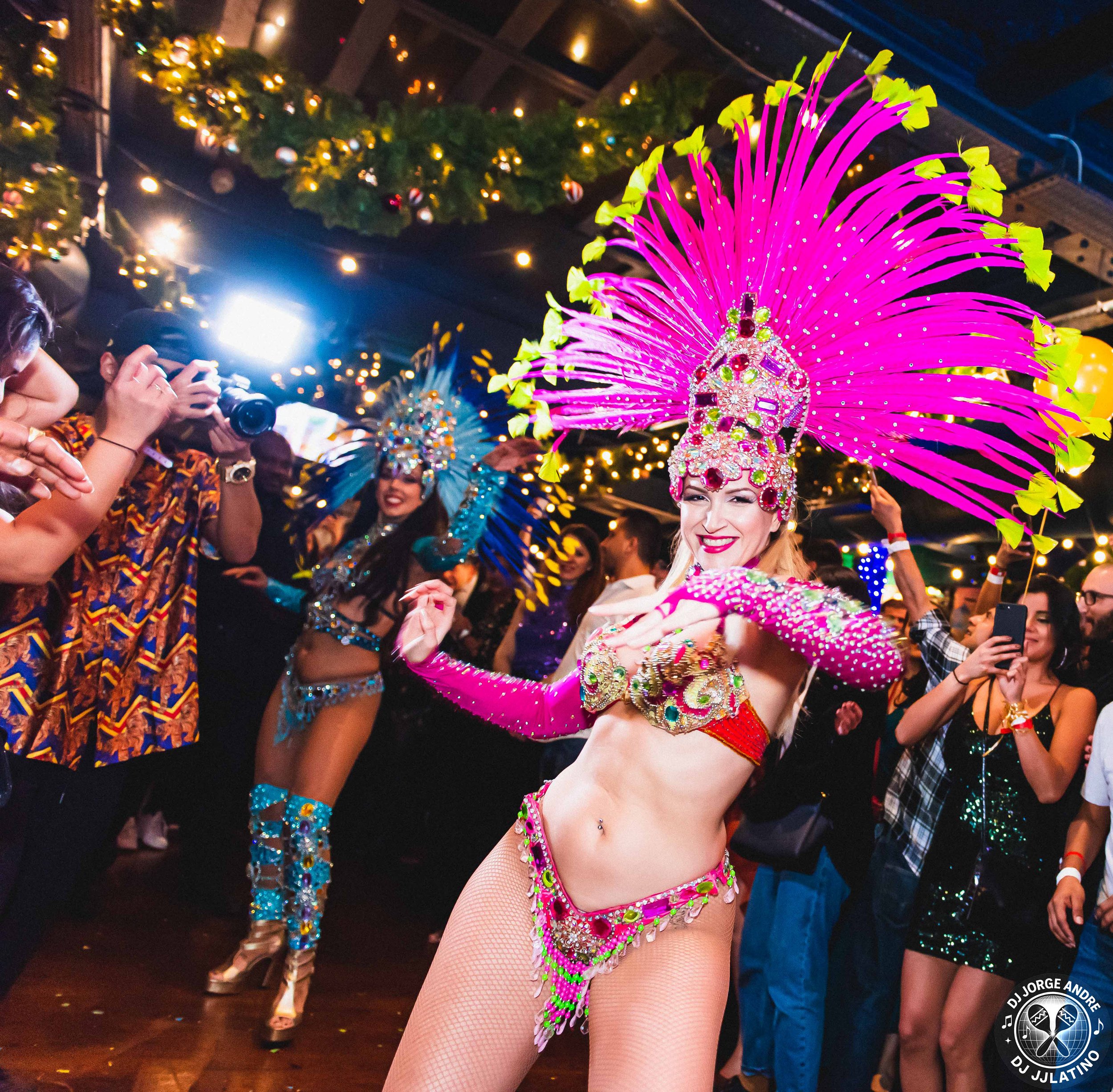 A dancer in a vibrant pink feathered headdress and elaborate costume performing at a lively party, surrounded by onlookers and other performers, with festive lighting and decorations in the background.