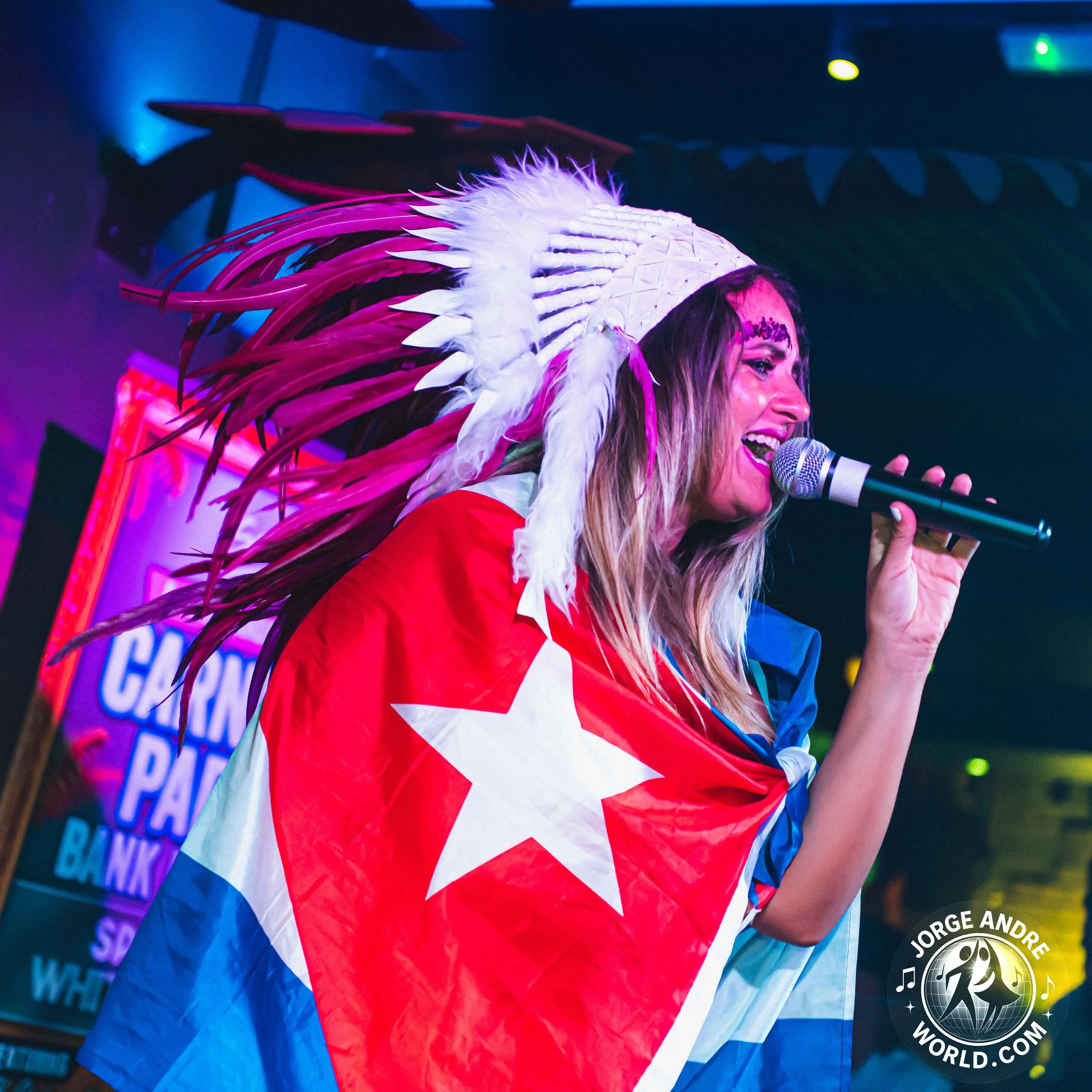 A woman singing into a microphone while wearing a large Native American headdress and a Cuban flag as a cape in a lively, colorful setting.