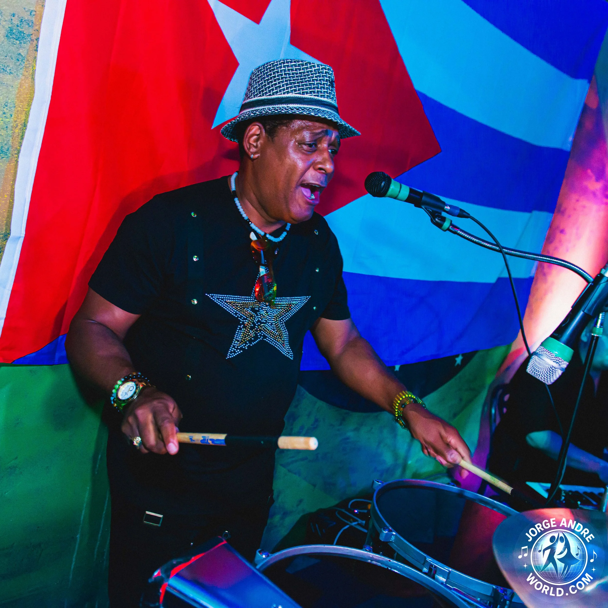 A man wearing a hat, star shirt, and jewelry is playing drums and singing into a microphone in front of a Cuban flag.