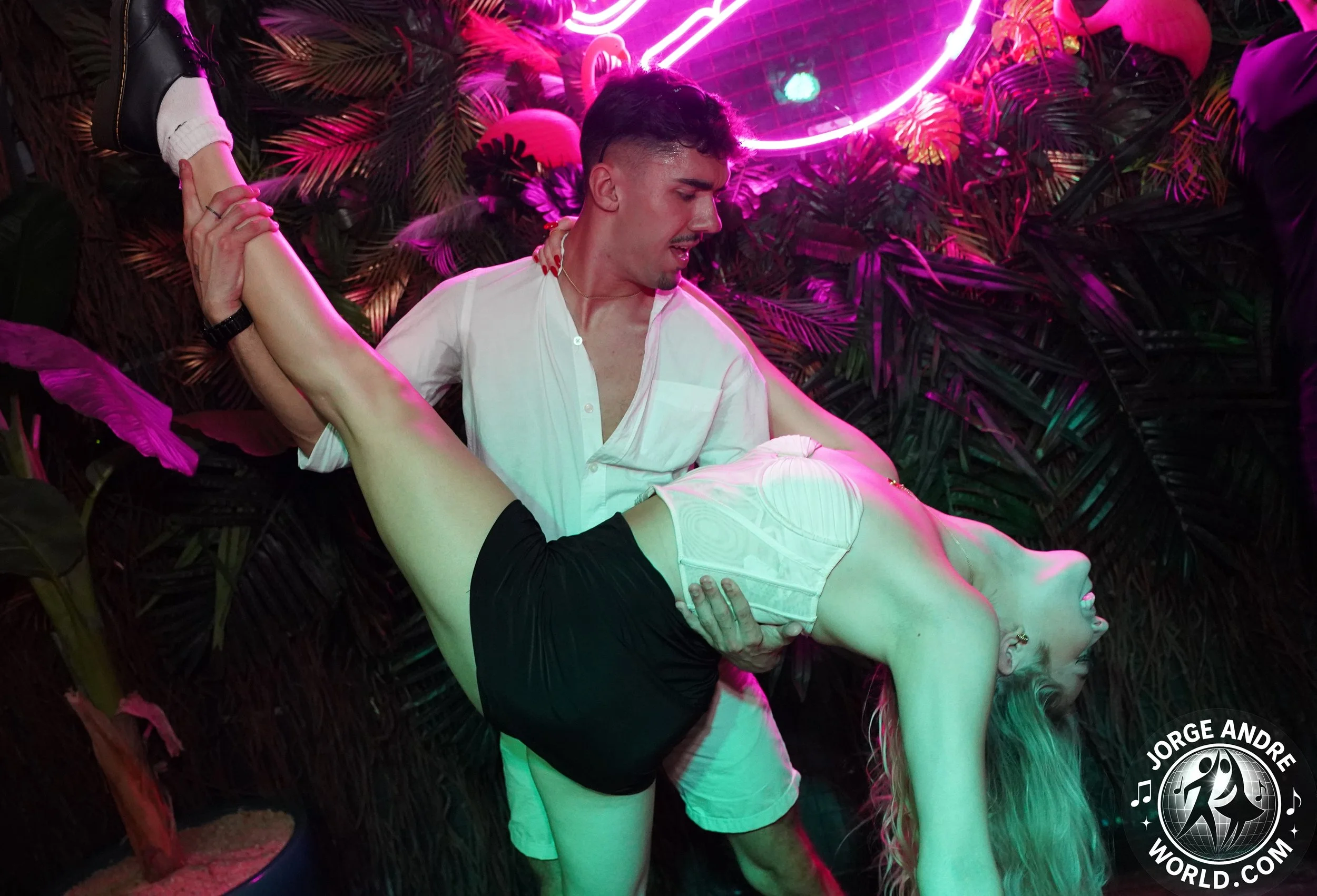 A man is holding a woman in a dance pose at a party with neon lights and tropical plants in the background.
