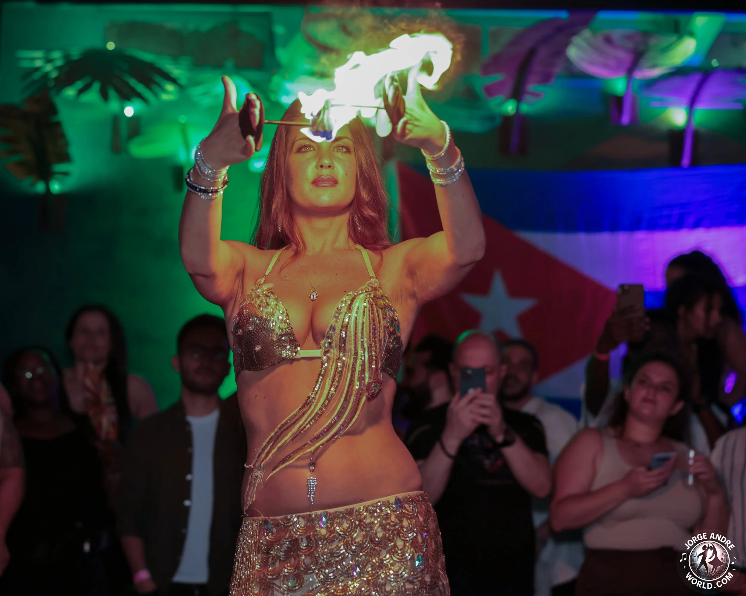 A woman performing a fire act with a flaming stick during a cultural event, with an audience watching and taking photos, and a flag in the background.