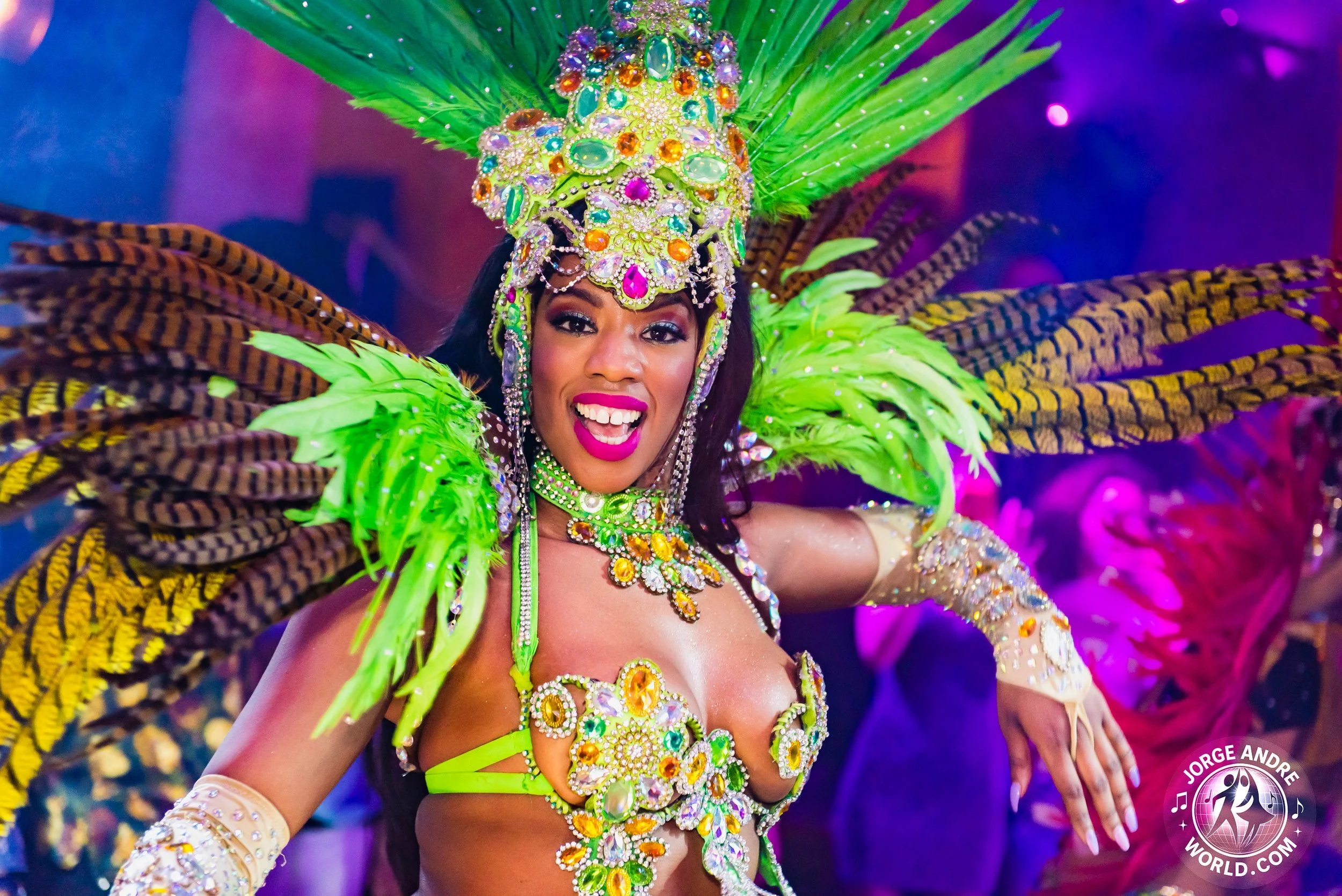 A woman in a colorful carnival costume with bright green and yellow feathers, decorated with rhinestones and jewelry, smiling and dancing at a celebration.