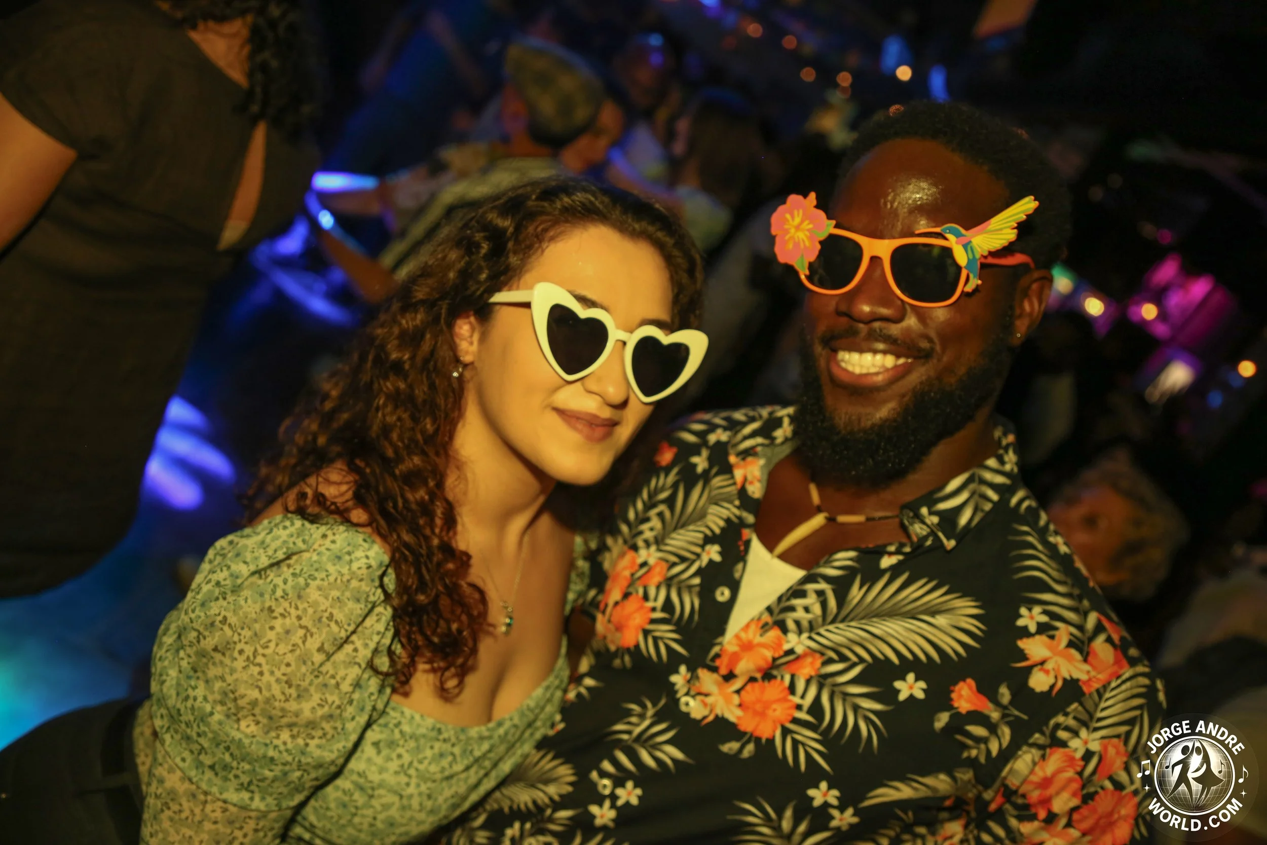 Two people smiling at a party wearing fun sunglasses; the woman has heart-shaped sunglasses and the man has Hummingbird sunglasses, both dressed in tropical-themed attire.