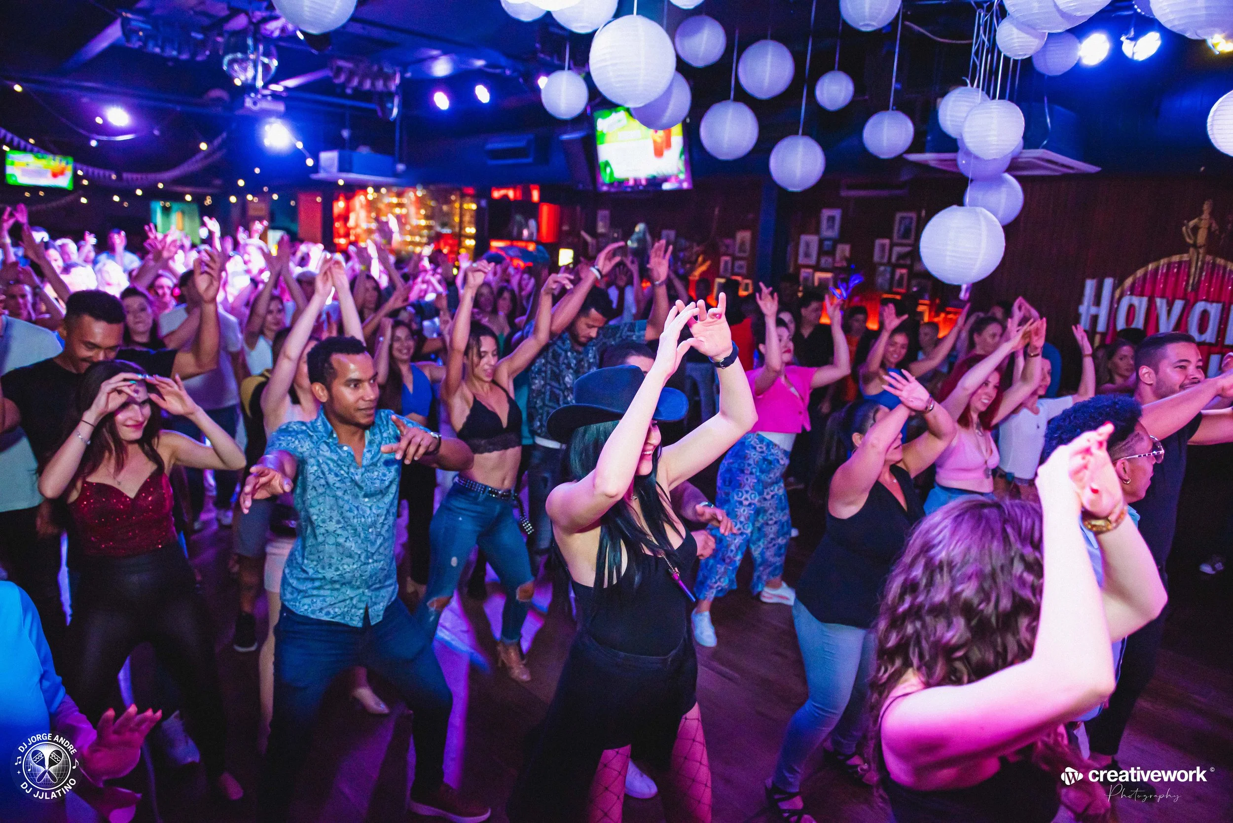 Crowd of people dancing at a lively indoor party with hanging paper lanterns and colorful lighting.