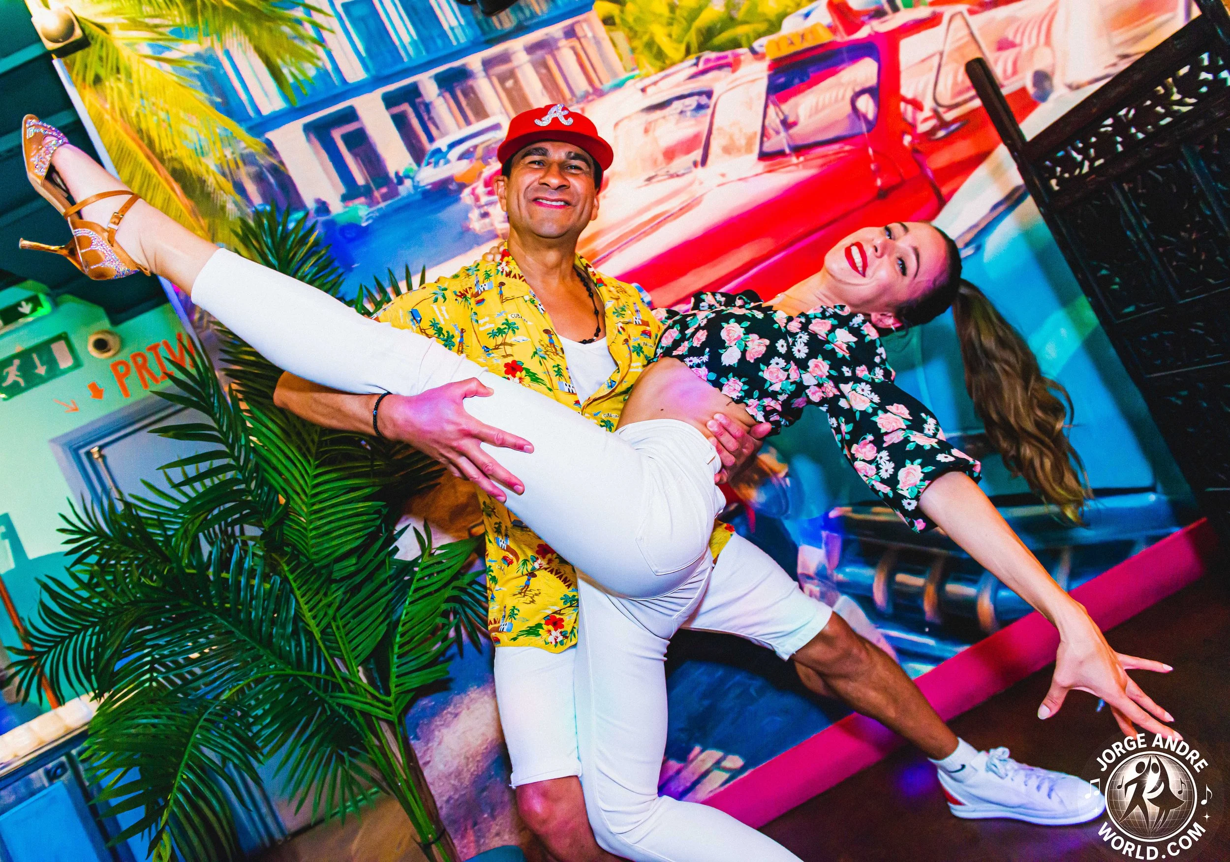 A man wearing a red cap, yellow Hawaiian shirt, and white shorts is holding a woman in a black floral top and white pants in a dance pose at a colorful indoor setting with a Miami backdrop and tropical plants.