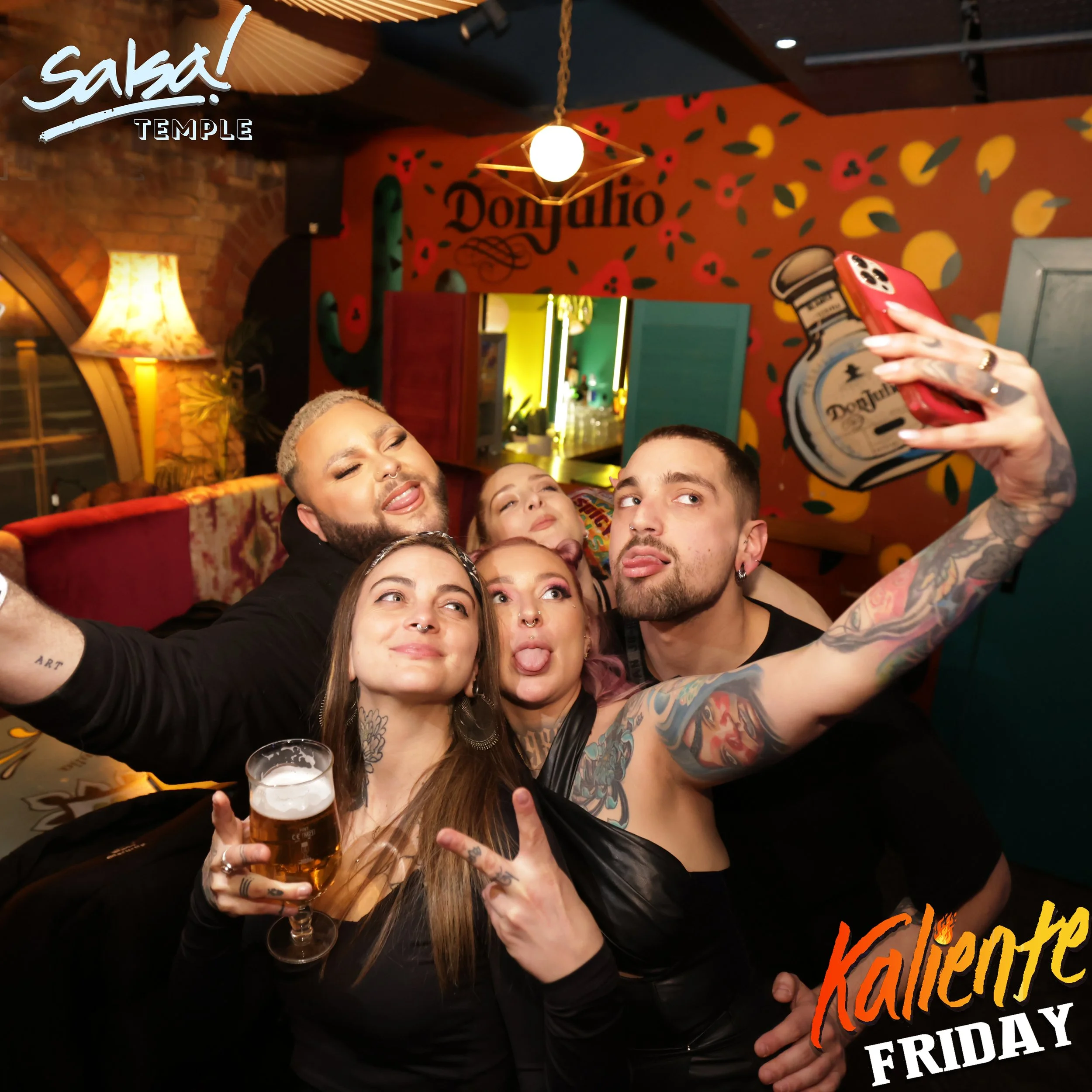 Group of friends taking a selfie at a lively restaurant or bar, some making peace signs and sticking out tongues, with colorful wall decorations and drinks in hand.
