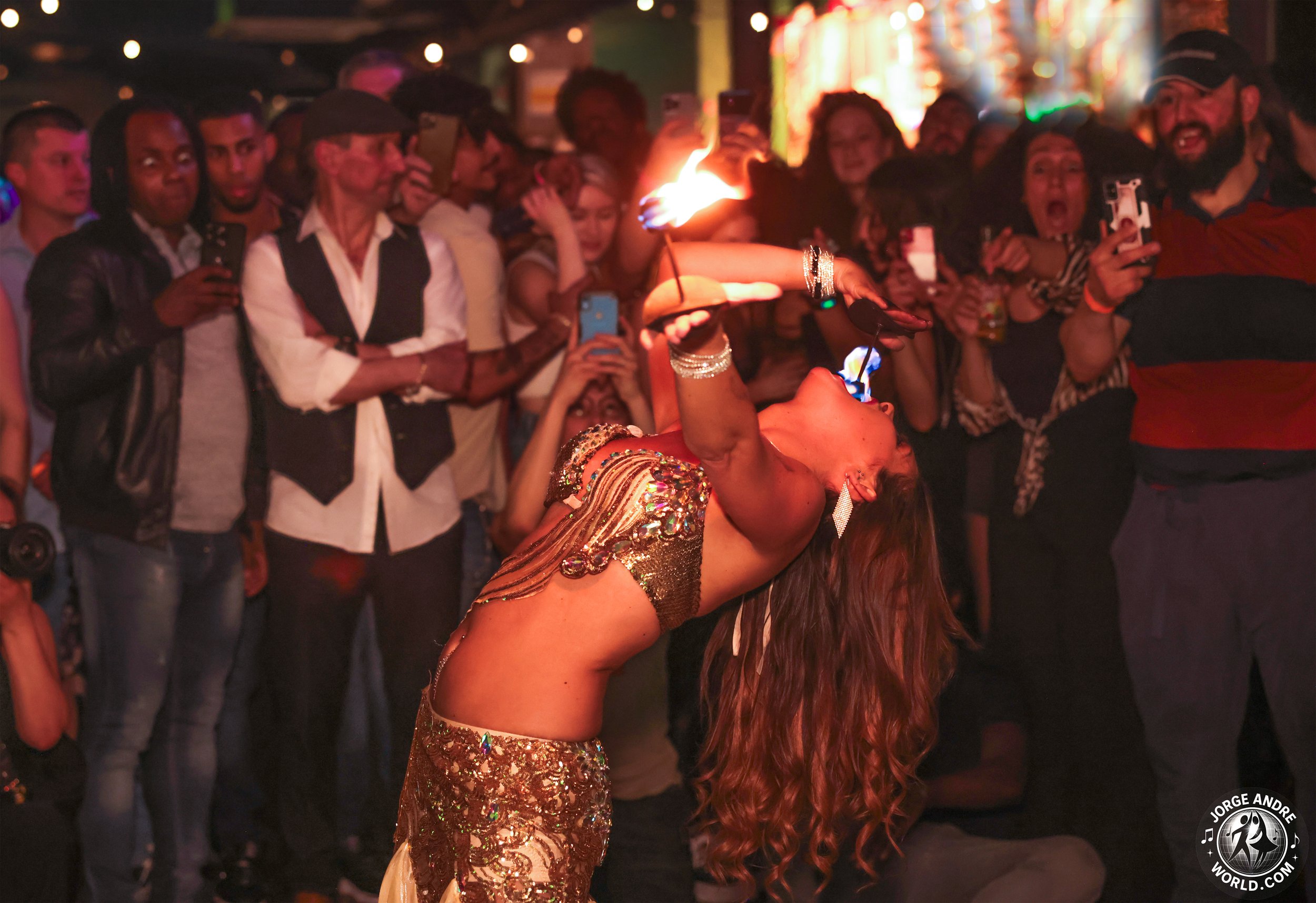 A woman in a sparkling gold dress performs a fire-breathing act in front of a crowd of onlookers at a nightlife event. She is leaning back with her mouth open and holding a lit torch, with flames coming out of her mouth. The bystanders are watching a