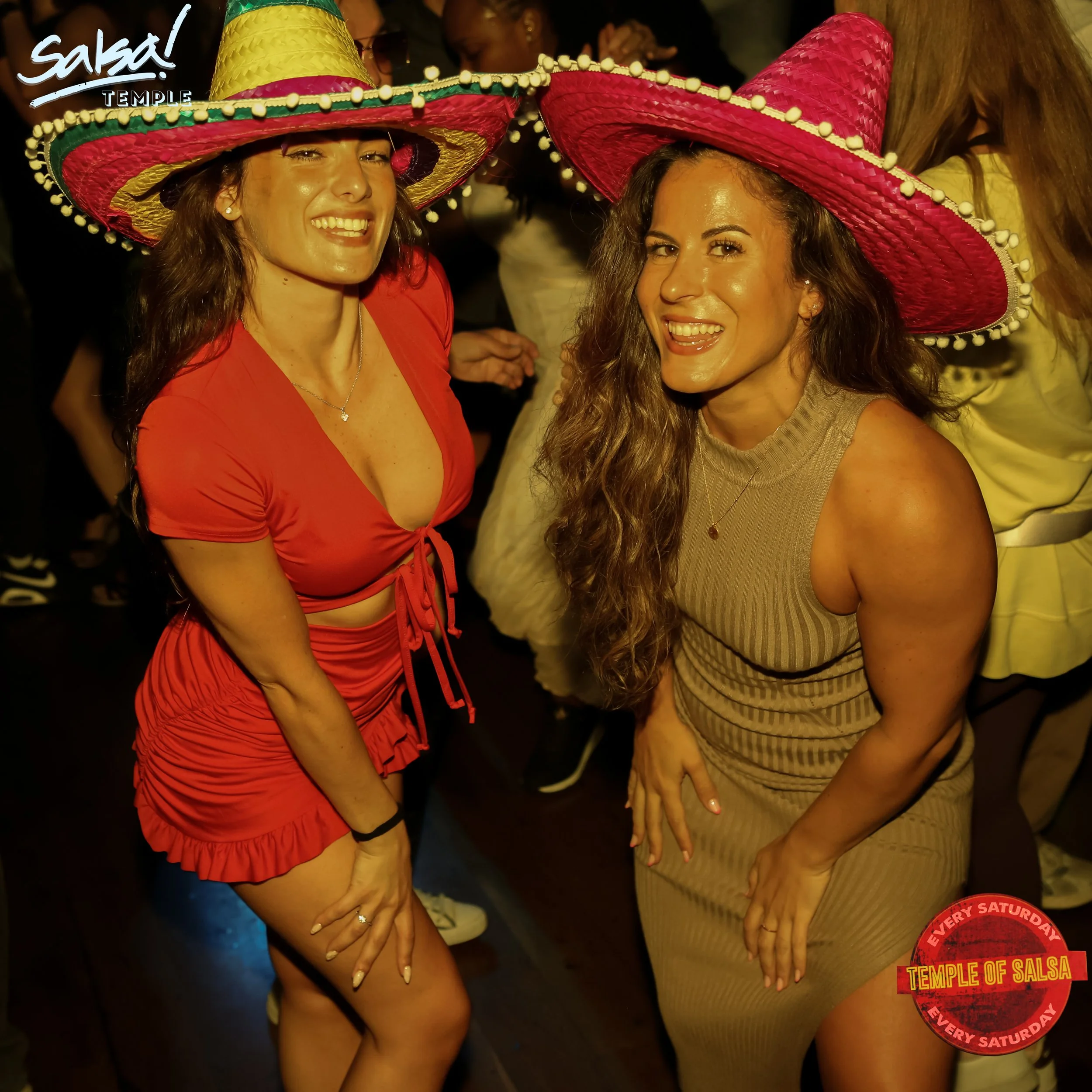 Two women wearing large pink and yellow sombreros, posing and smiling at a salsa dance event, with a 'Temple of Salsa' logo in the bottom right corner.