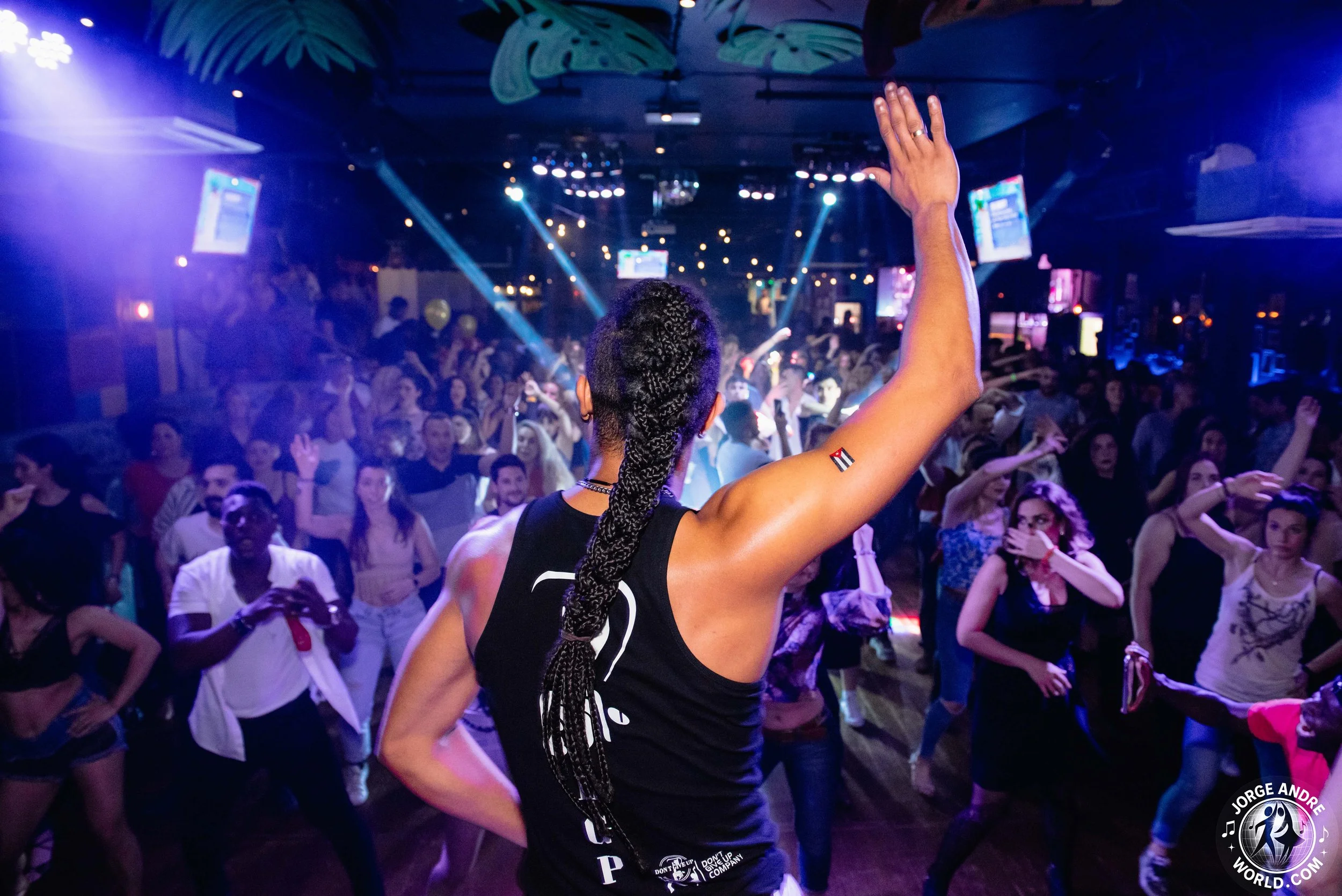 A woman with braided hair and a temporary tattoo on her arm leading a dance or exercise class in a crowded club or dance venue with colorful lighting and a lively crowd.
