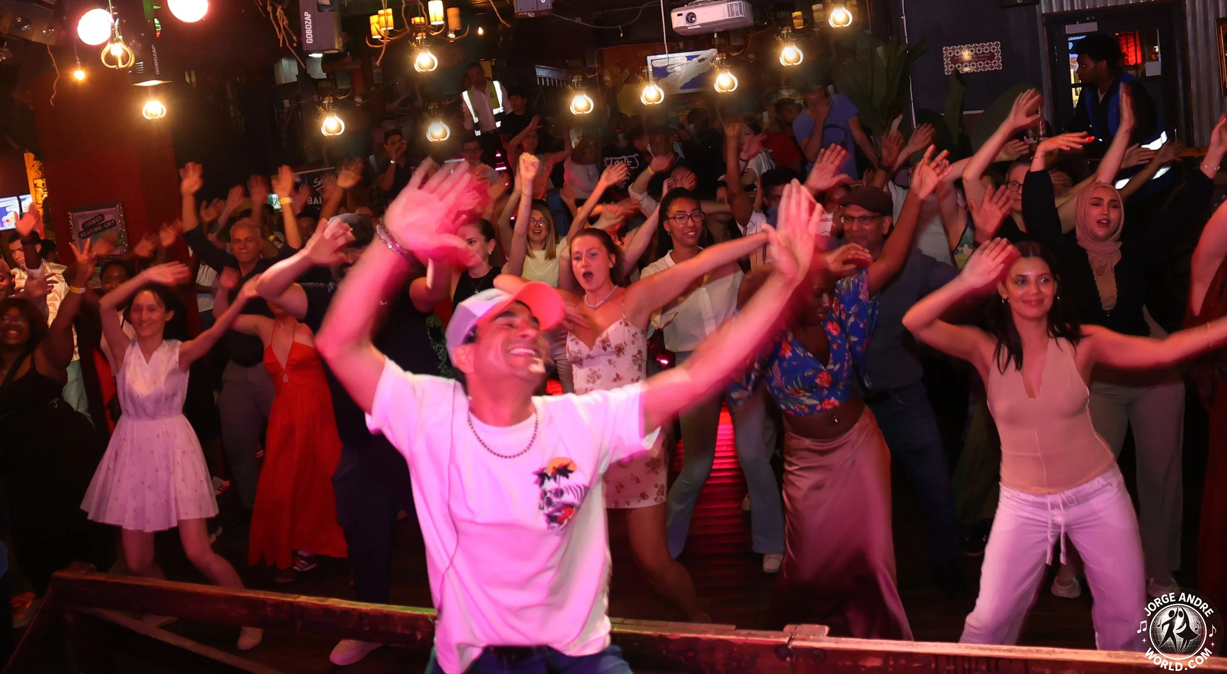 People dancing and having fun in a lively indoor nightclub or party setting, with colorful lighting and some individuals smiling and raising their hands.