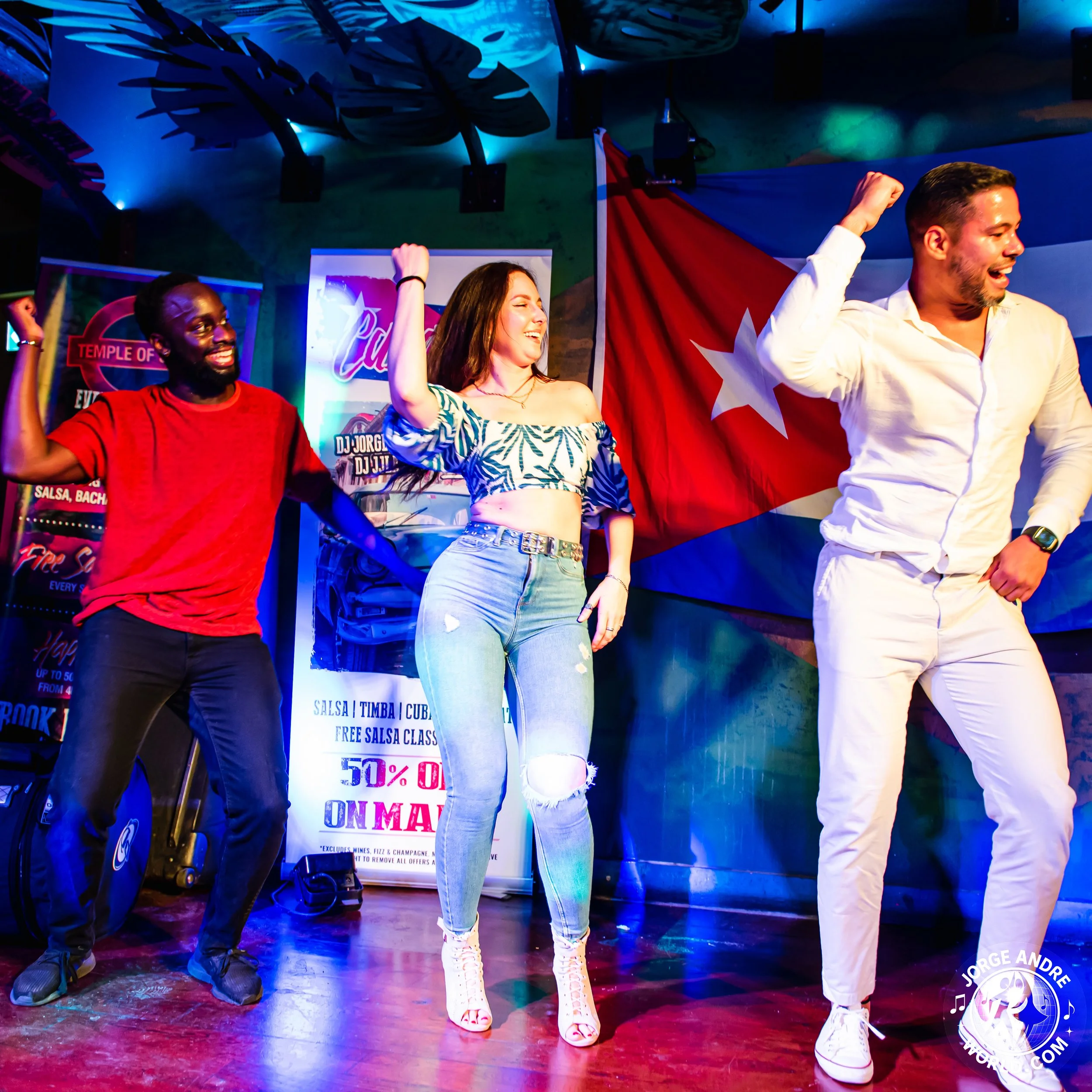Three people dancing and enjoying music in a lively setting with a Cuban flag in the background, colorful lighting, and a promotional sign for salsa classes.