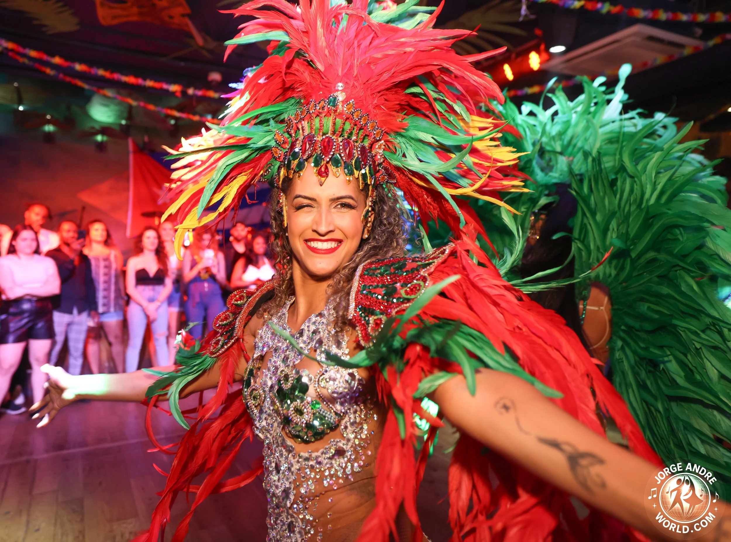 A woman samba dancer  dressed in a colorful and ornate costume, wearing a large headdress made of red, green, and yellow feathers, smiling and posing at a lively event with a group of people in the background.
