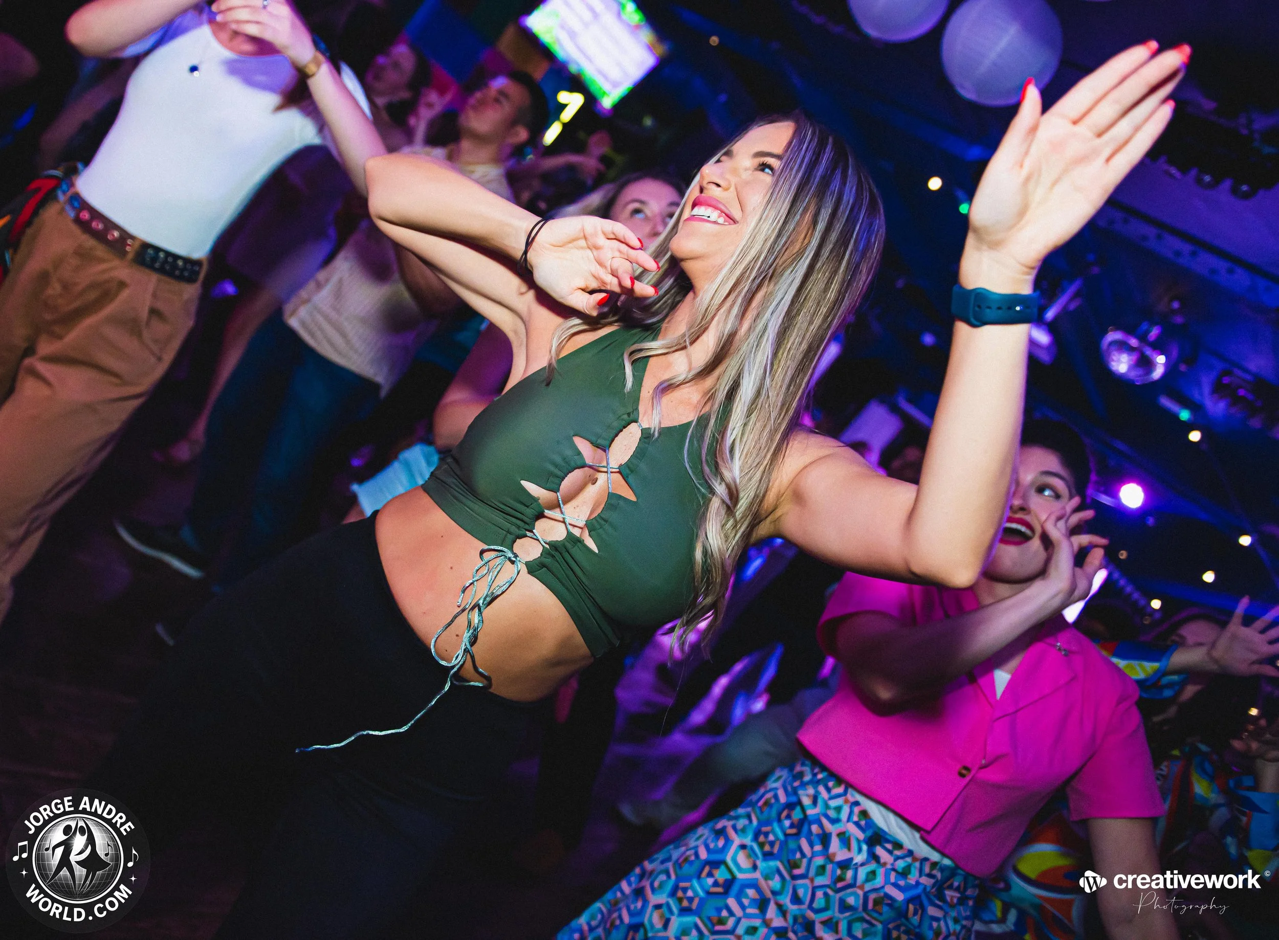 Young woman dancing at a party with colorful lighting, wearing a green cropped top and black pants, smiling with her eyes closed, surrounded by other partygoers.