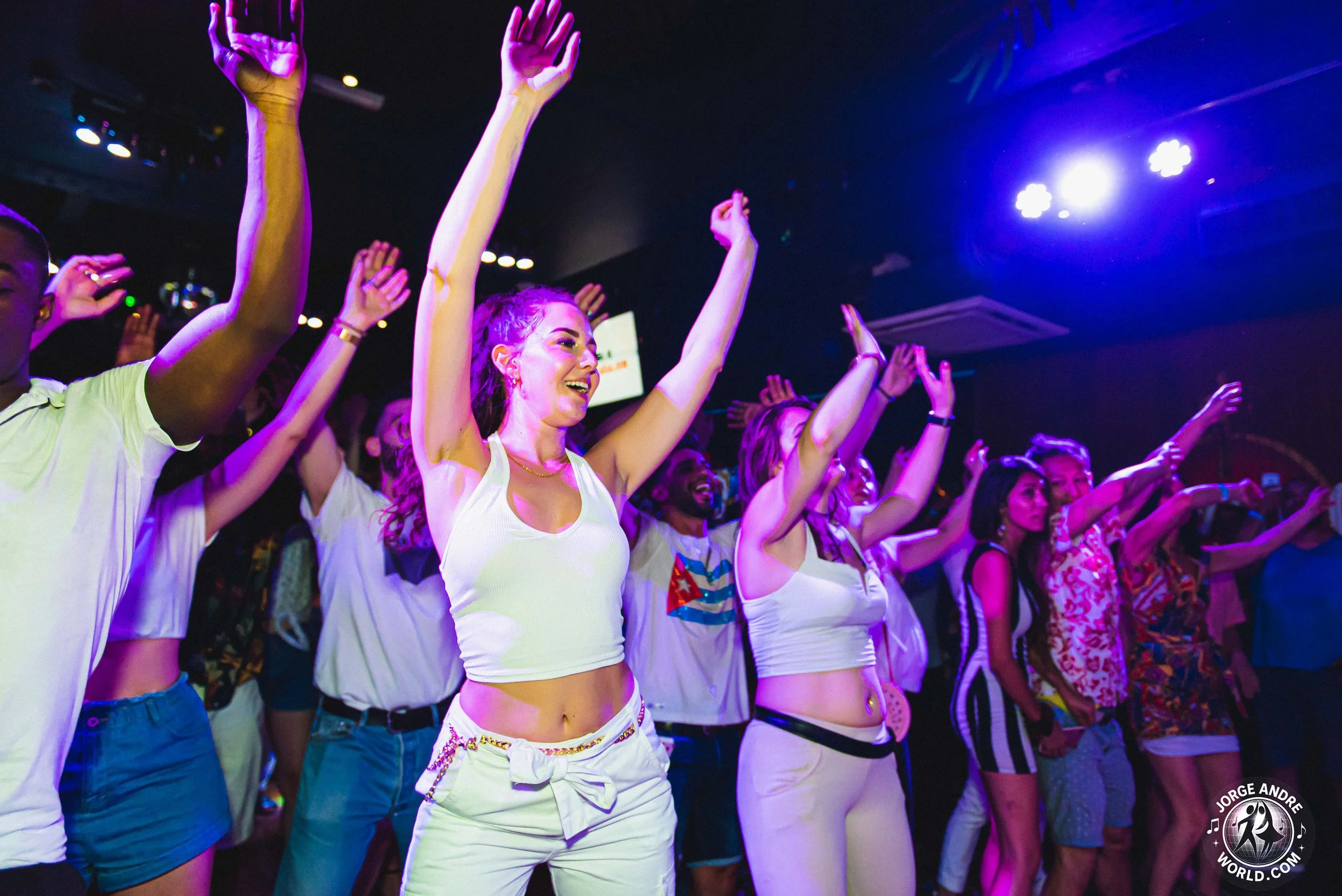 People dancing with hands raised in a club or concert setting, colored lighting, vibrant atmosphere.