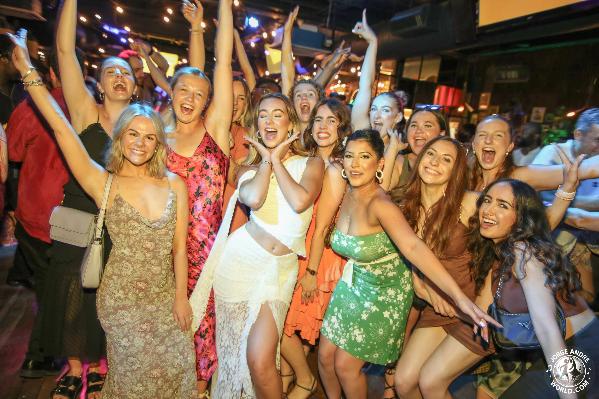 A group of women at a lively party, smiling and dancing with their arms raised in a nightclub or party setting with colorful lights.